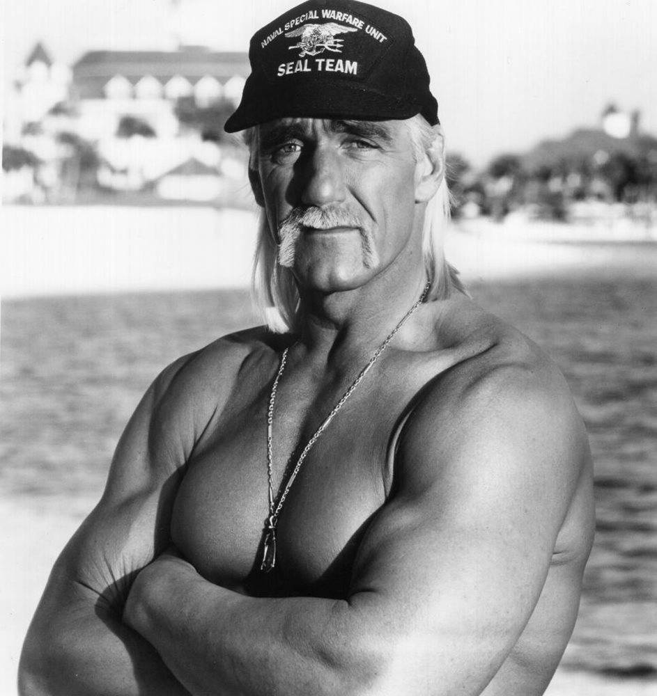 經典灰階 Hulk Hogan 壁紙 — 一個肌肉發達、留著金色長髮和鬍鬚的男人，赤裸上身站在沙灘上，戴著SEAL TEAM帽子和項鍊。