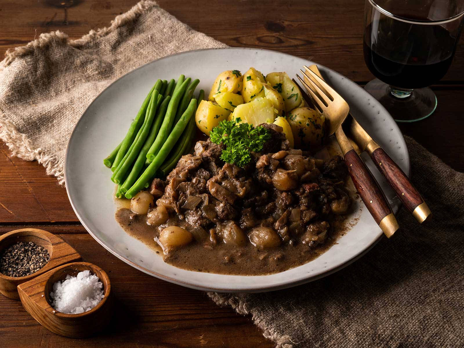 經典牛肉布丁醬配馬鈴薯與豆類 壁紙 — 一盤鄉村風味的燉牛肉配馬鈴薯和青豆，飾以歐芹，放在木桌上，旁邊有一杯紅酒。