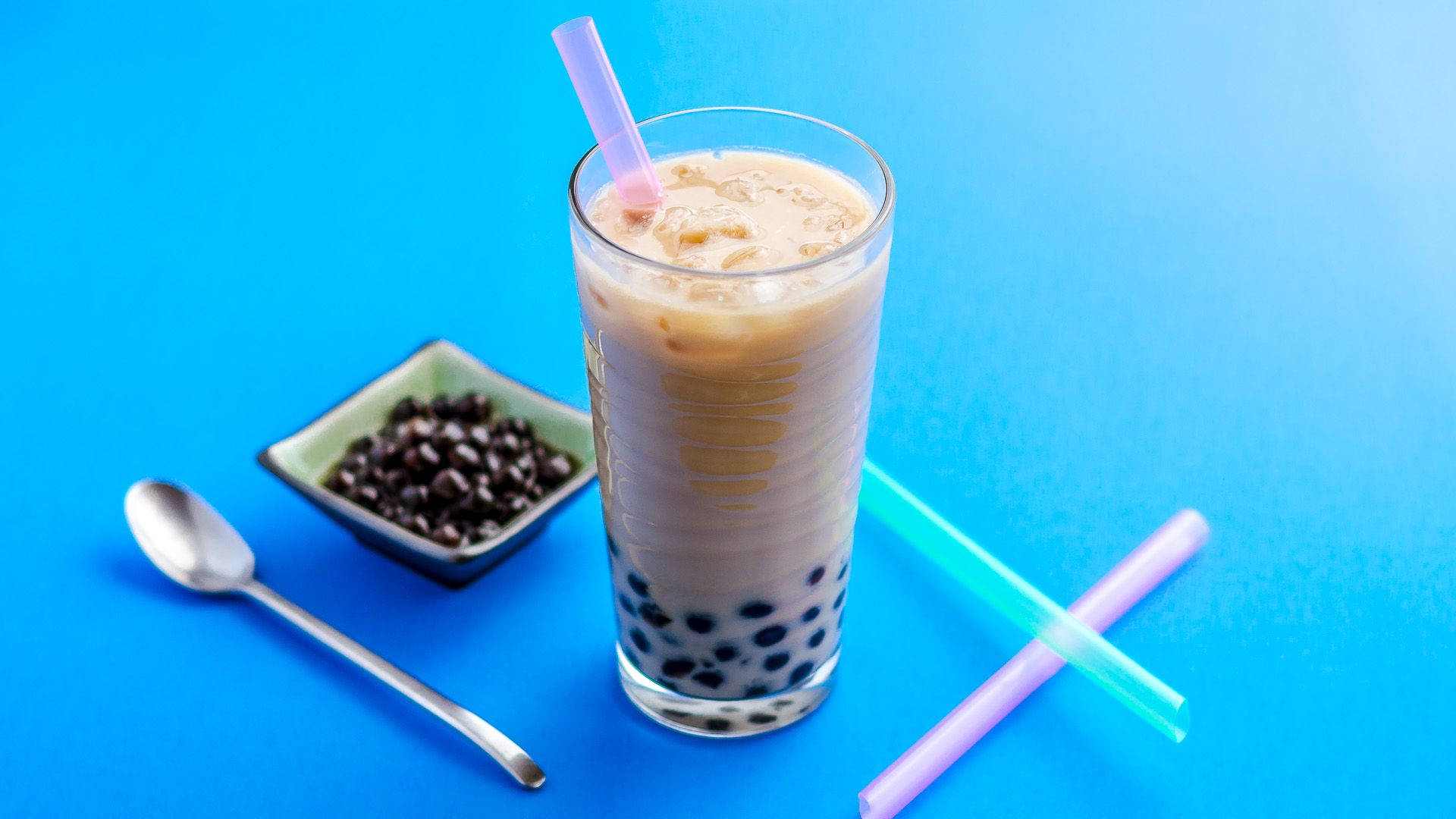 經典珍珠奶茶 壁紙 — 一杯高身的冰珍珠奶茶，配有木薯珍珠和粉紅色吸管，旁邊放著一小碟珍珠和彩色吸管，背景為明亮的藍色。