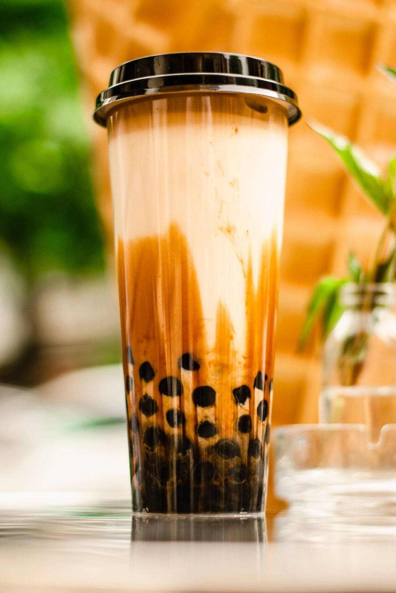 經典琥珀奶茶 壁紙 — 一杯高身透明杯裝著 creamy 焦糖泡泡茶，配以黑色木薯珍珠，放在反光表面上，背景溫暖模糊。