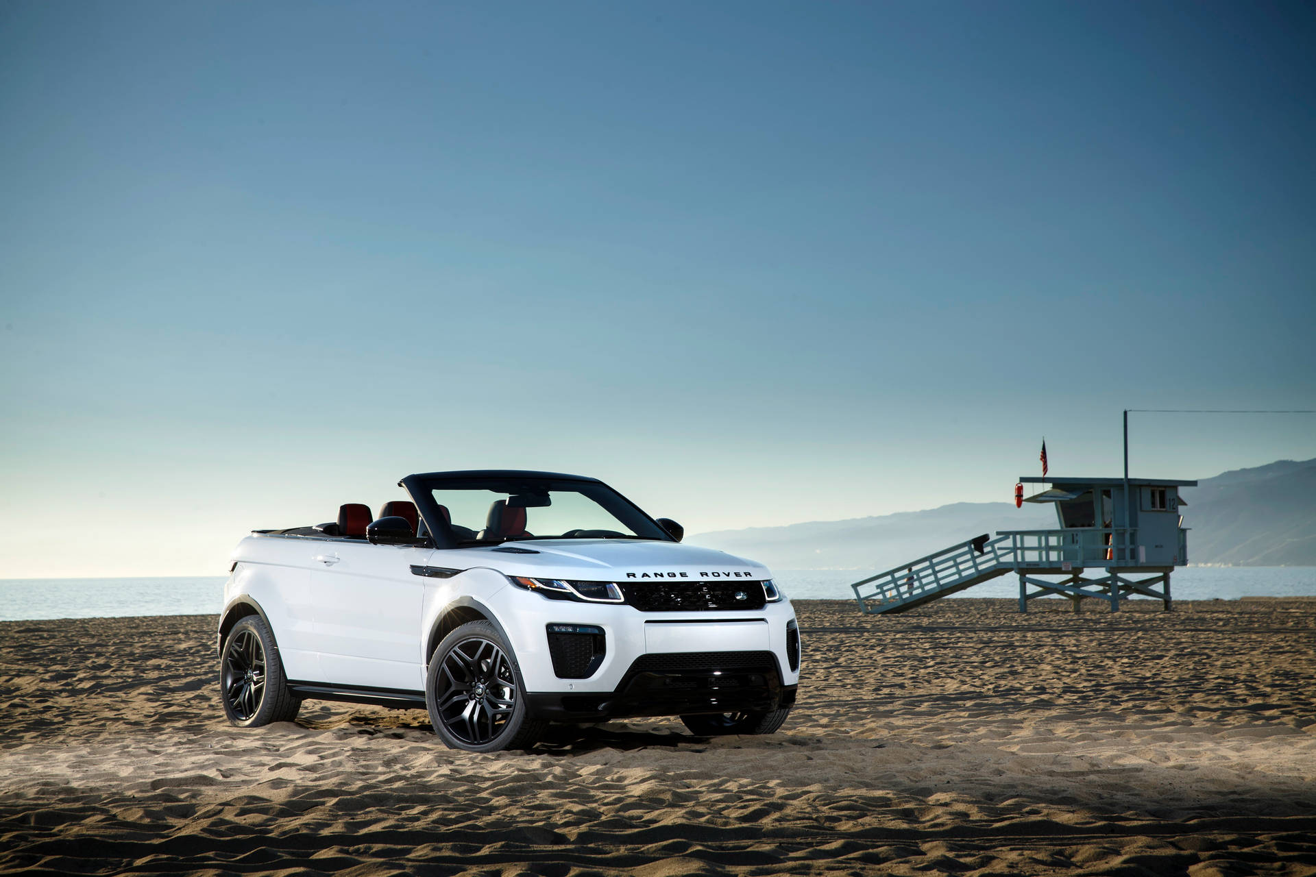 白色 Range Rover Evoque Convertible 停泊在沙灘上，靠近救生員瞭望塔，夕陽西下。