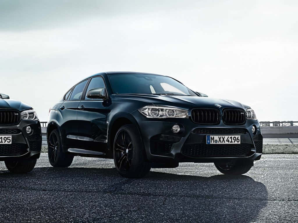 經典的 BMW X6 M 是一款強勁性能車 壁紙 — 兩輛時尚的黑色BMW SUV停泊在瀝青路面上，天空多雲，帶有戲劇性的陰影。