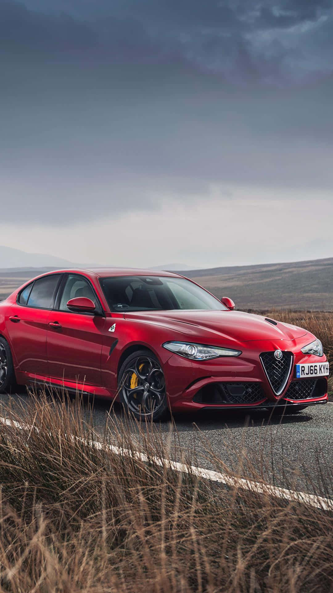 經典的Alfa Romeo 壁紙 — 一輛鮮紅色的愛快羅密歐Giulia Quadrifoglio在陰沉多雲的天空下沿著蜿蜒的山路飛馳。