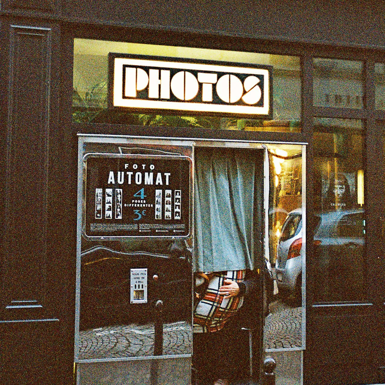 經典相機是派對的完美點綴 壁紙 — 一個復古照相亭，帶有發光的「PHOTOS」標誌，裡面有一個穿著格子衣服的人拿著相機。