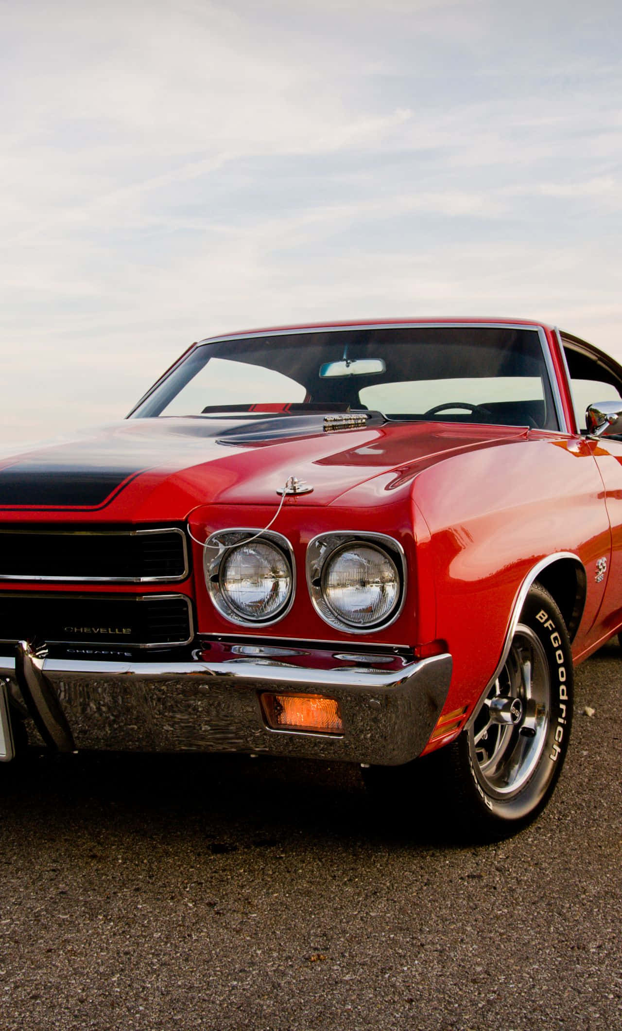 經典紅色 Chevrolet Chevelle 車型 iPhone 壁紙 — 一架光面紅色經典雪佛蘭Chevelle肌肉車，帶有黑色引擎蓋條紋，停泊在陰天天空下的瀝青路面上。