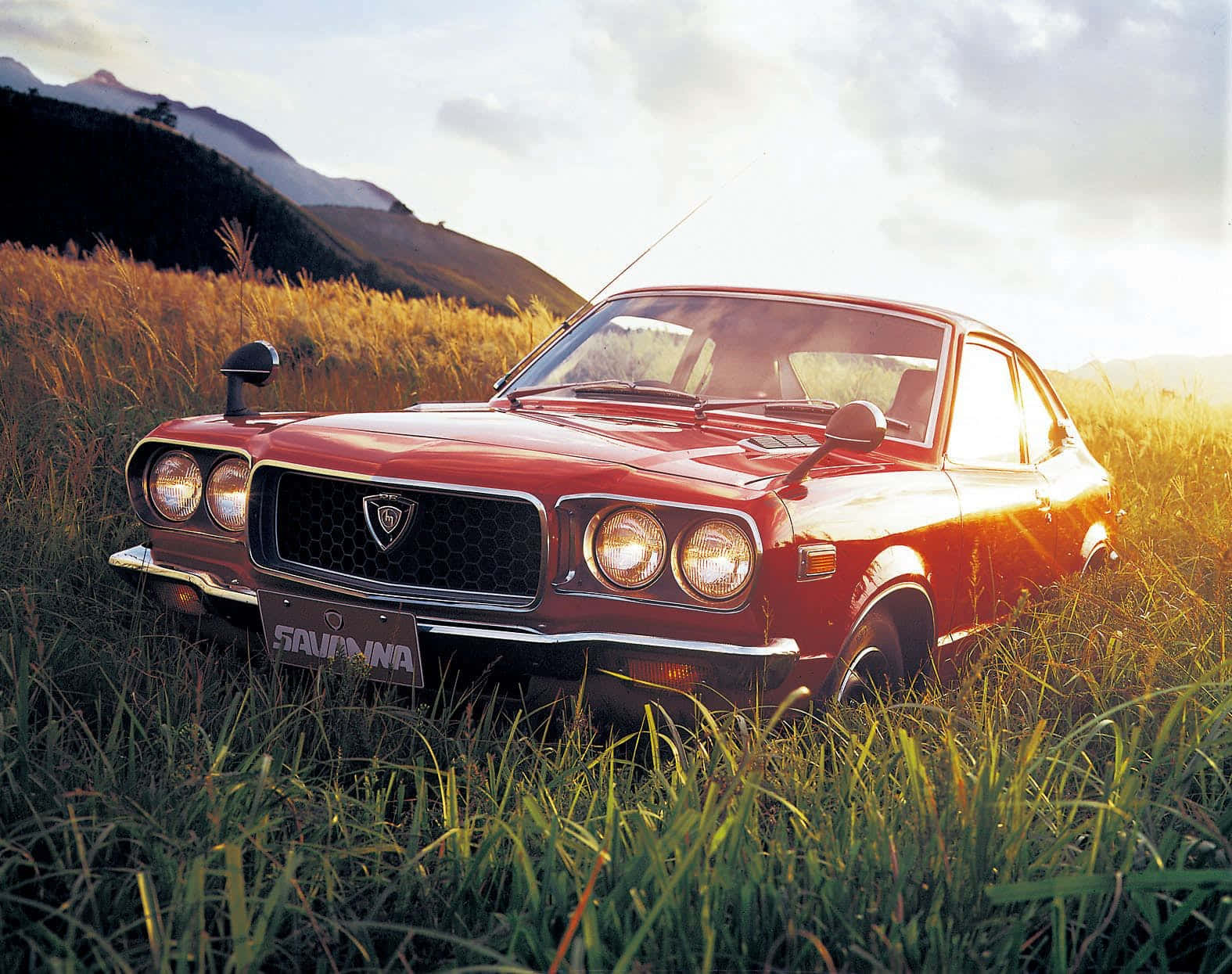 Mazda Rx-3 桌布 壁紙 — 經典紅色 Datsun SAVANNA 雙門跑車在金色夕陽下閃耀，停放在高草叢生的田野中，背後是起伏的丘陵。