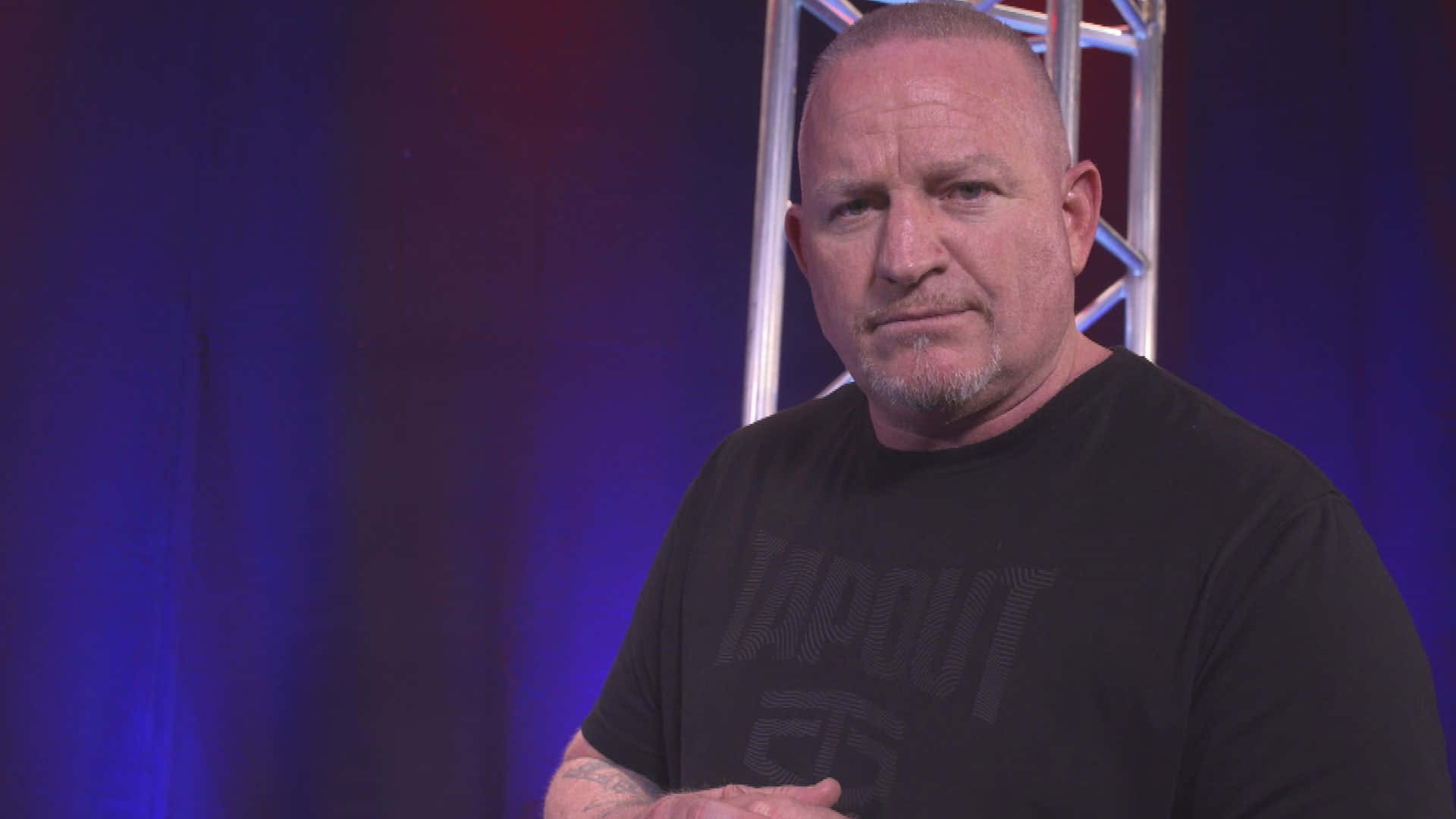 經典美國摔角手Road Dogg 壁紙 — 一個光頭留山羊鬍的男人穿著黑色T恤，站在藍色燈光背景前，身後有金屬桁架結構。