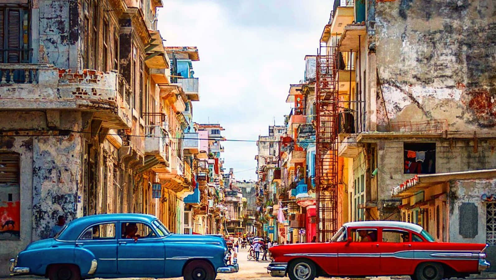 Cuba 桌布 壁紙 — 經典老爺車沿著陽光明媚的古巴街道行駛，兩旁是風化的粉彩建築，設有陽台和防火梯。