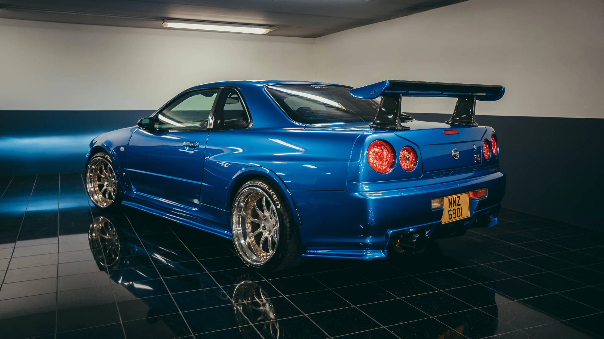 經典藍色 R34 GT-R 車款 壁紙 — 一輛光澤藍色的日產Skyline R34跑車，帶有大型後擾流板，在光線昏暗、舖有反光黑色瓷磚地板的車庫中展示。