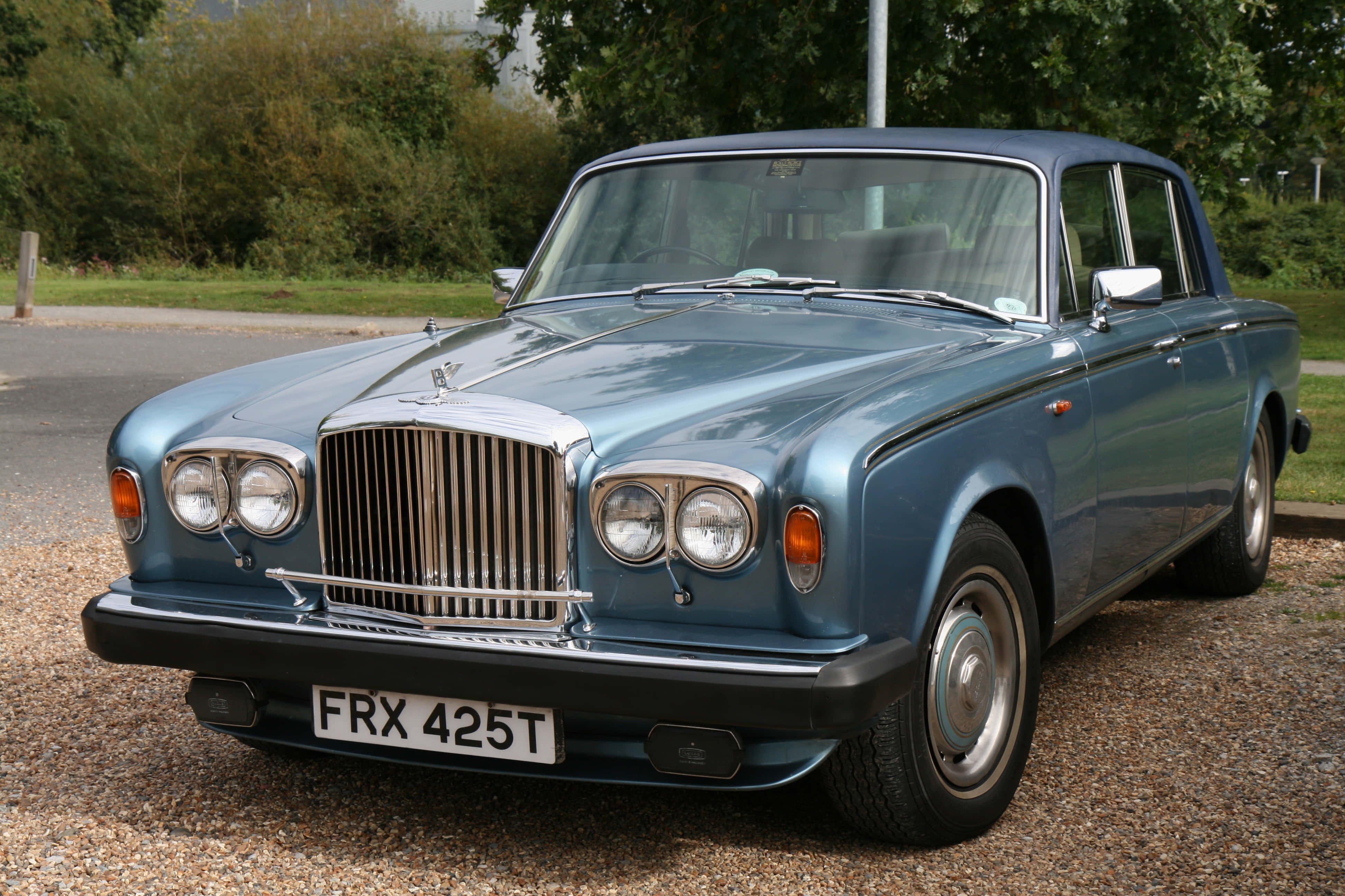 Bentley T2 桌布 壁紙 — 經典藍色賓利S3房車在日光下閃耀，停泊在碎石上，旁邊有綠色樹葉。