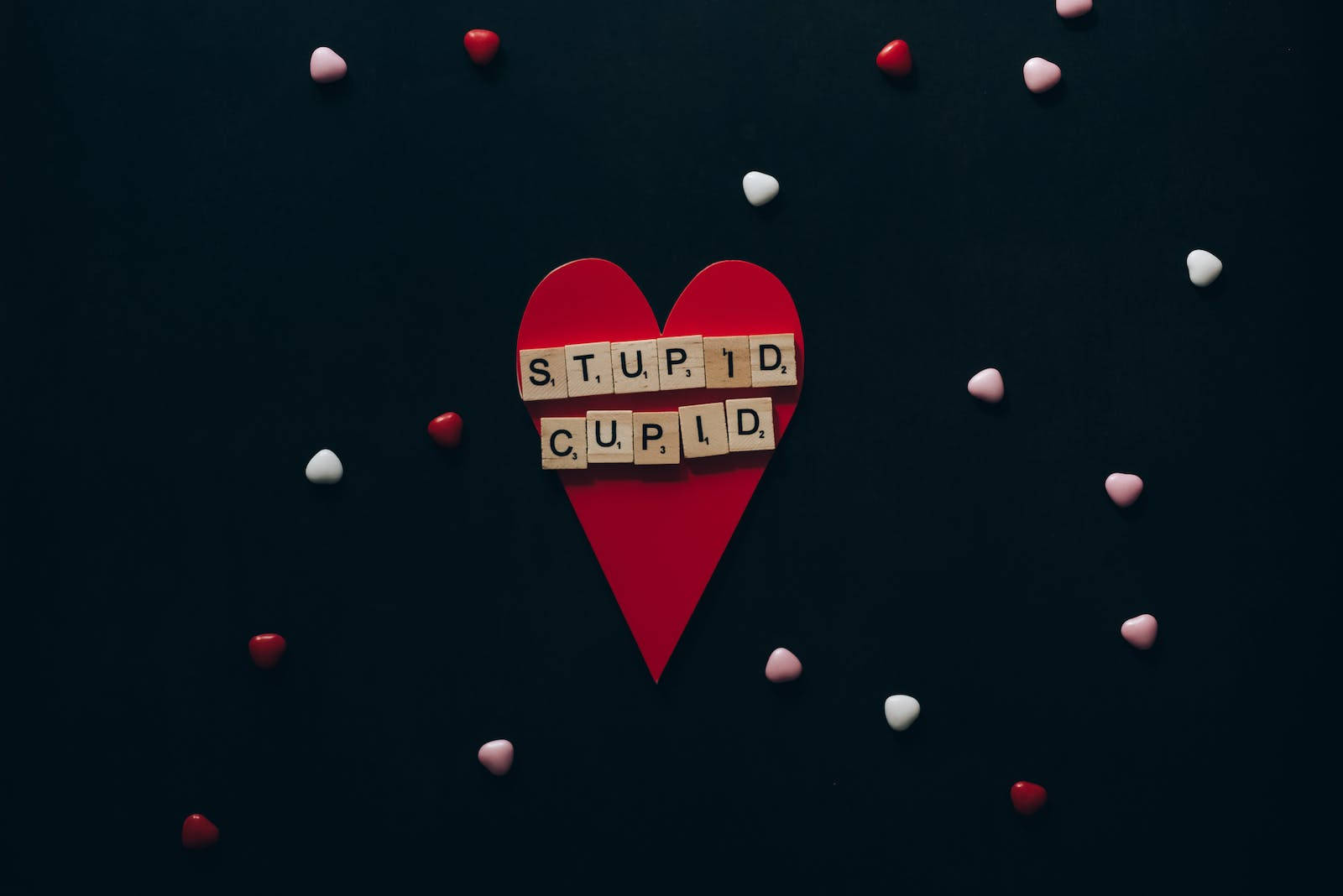 經典蠢丘比特壁紙 壁紙 — 一個紅色紙心，上面有木製拼字板拼出「STUPID CUPID」，周圍散落著紅色和白色的心形，背景為深色。