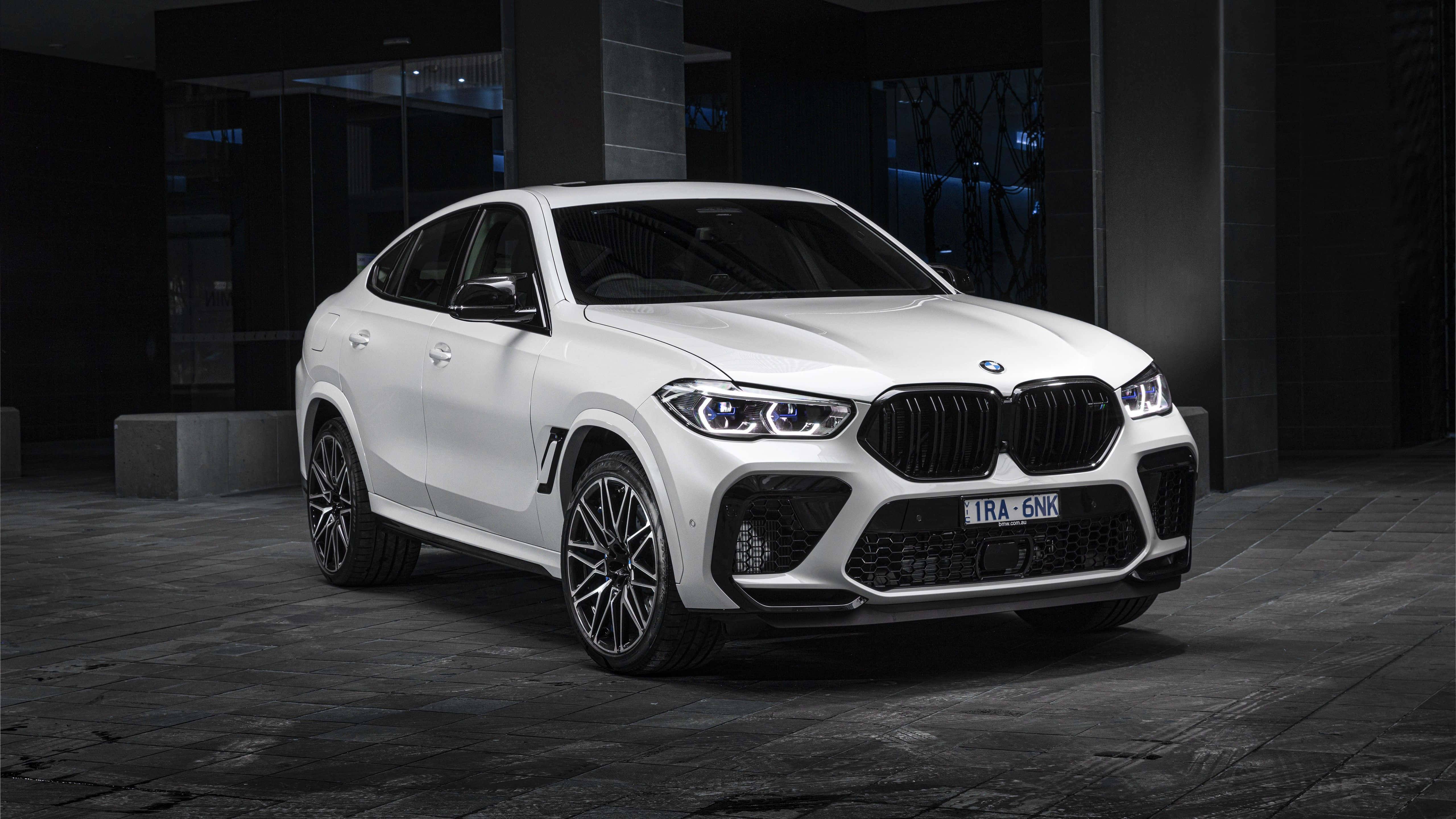 經典賽車血統的豪華駕馭 — BMW X6 M 壁紙 — 一輛時尚的白色BMW X6 M停放在現代建築下，車頭燈閃爍，周圍環境昏暗。