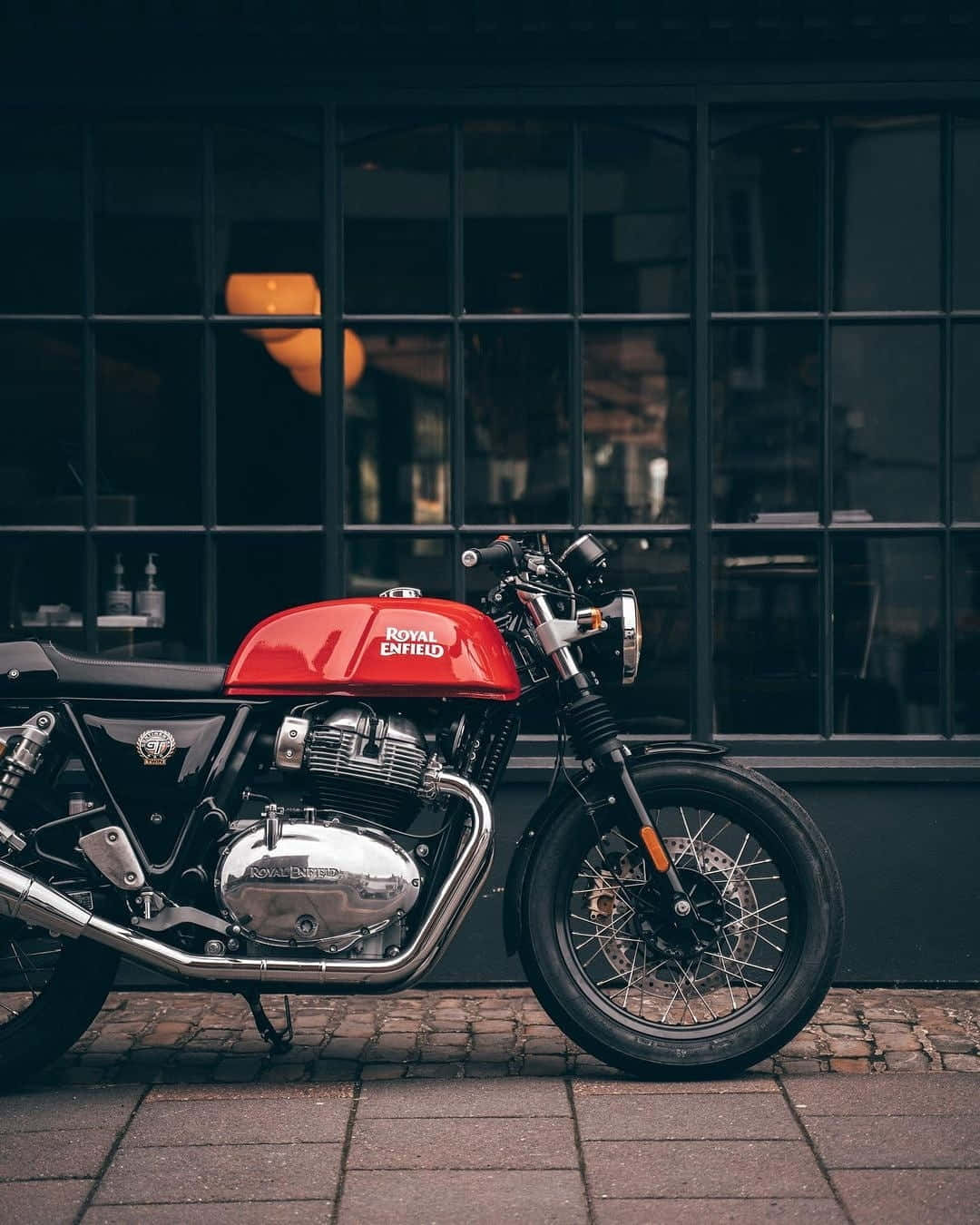 紅色 Royal Enfield 電單車夜間停泊在鵝卵石街道上，靠近玻璃建築。