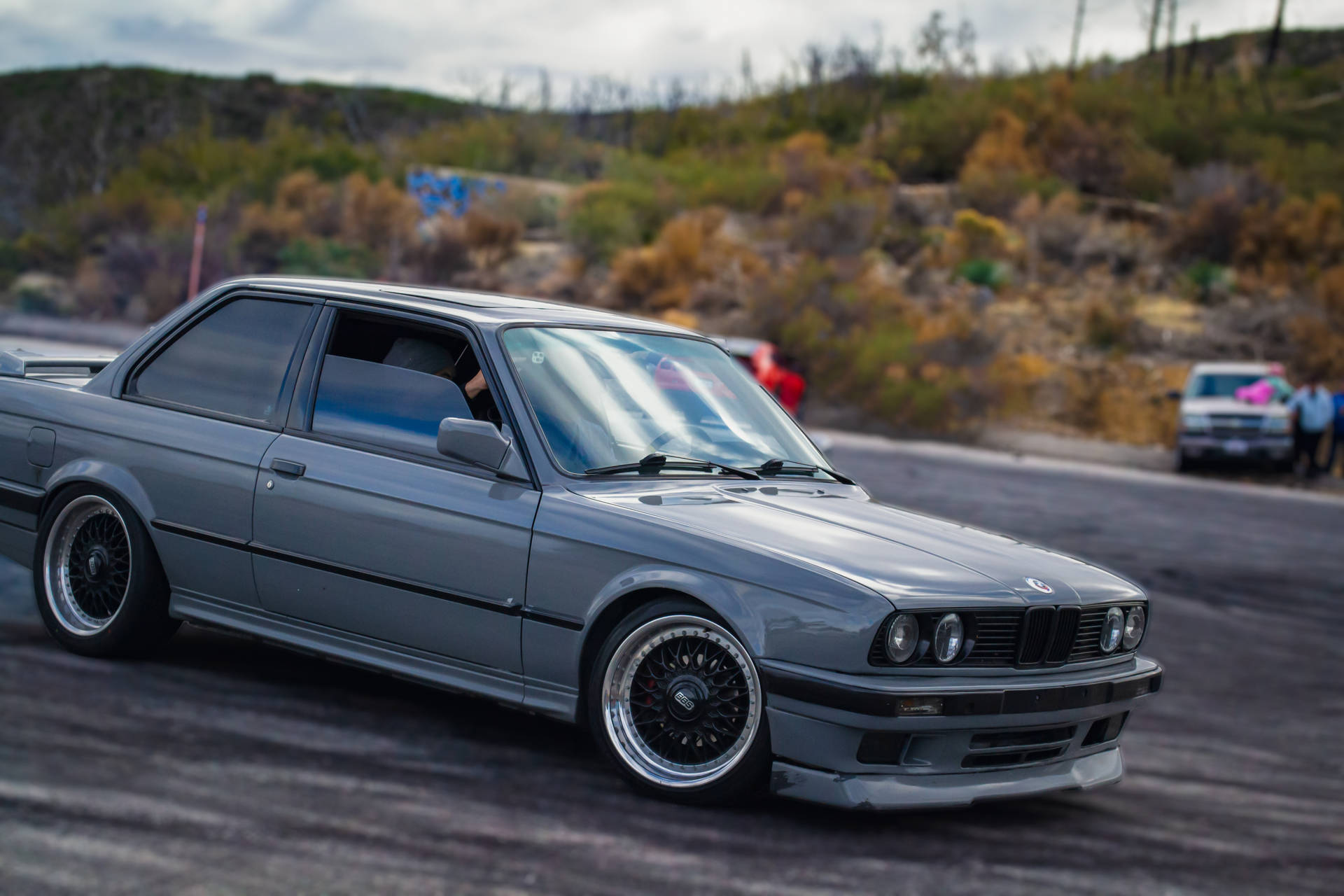 灰色BMW E30在賽道上飄移，後面有秋色山丘。