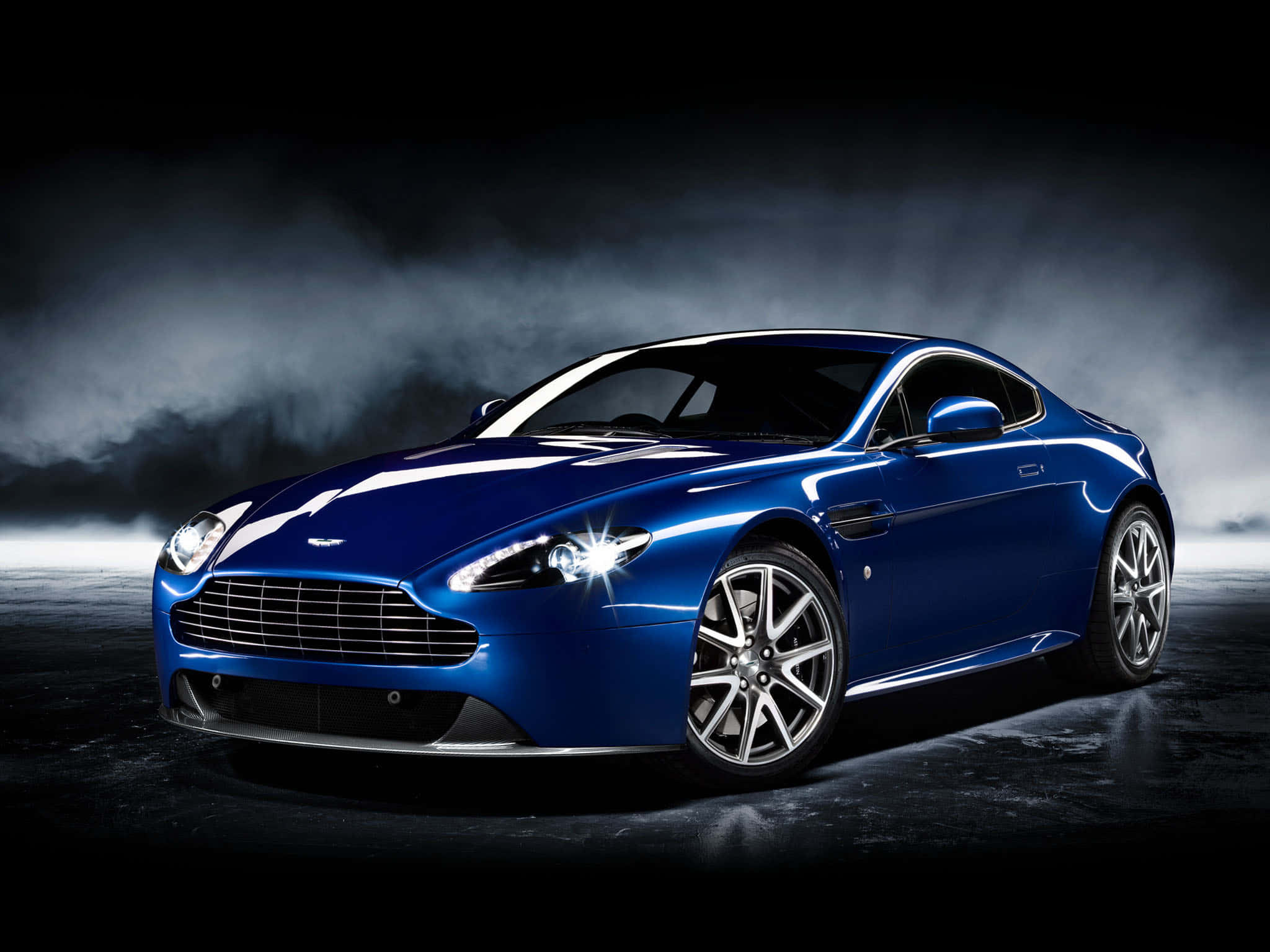 經典阿斯頓·馬丁配以鎂銀色更顯優雅 壁紙 — 一輛時尚的藍色Aston Martin跑車在戲劇性的工作室燈光下，在煙霧瀰漫的黑暗背景中閃閃發光。
