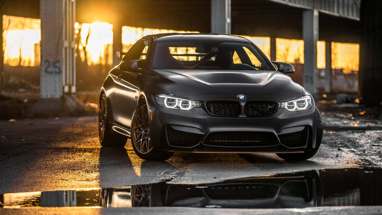 經典風格BMW，光澤黑漆車身 壁紙 — 一輛啞光黑色BMW M4雙門轎跑車在金色夕陽下閃耀，其倒影在工業支柱下濕混凝土上的水坑中閃爍。