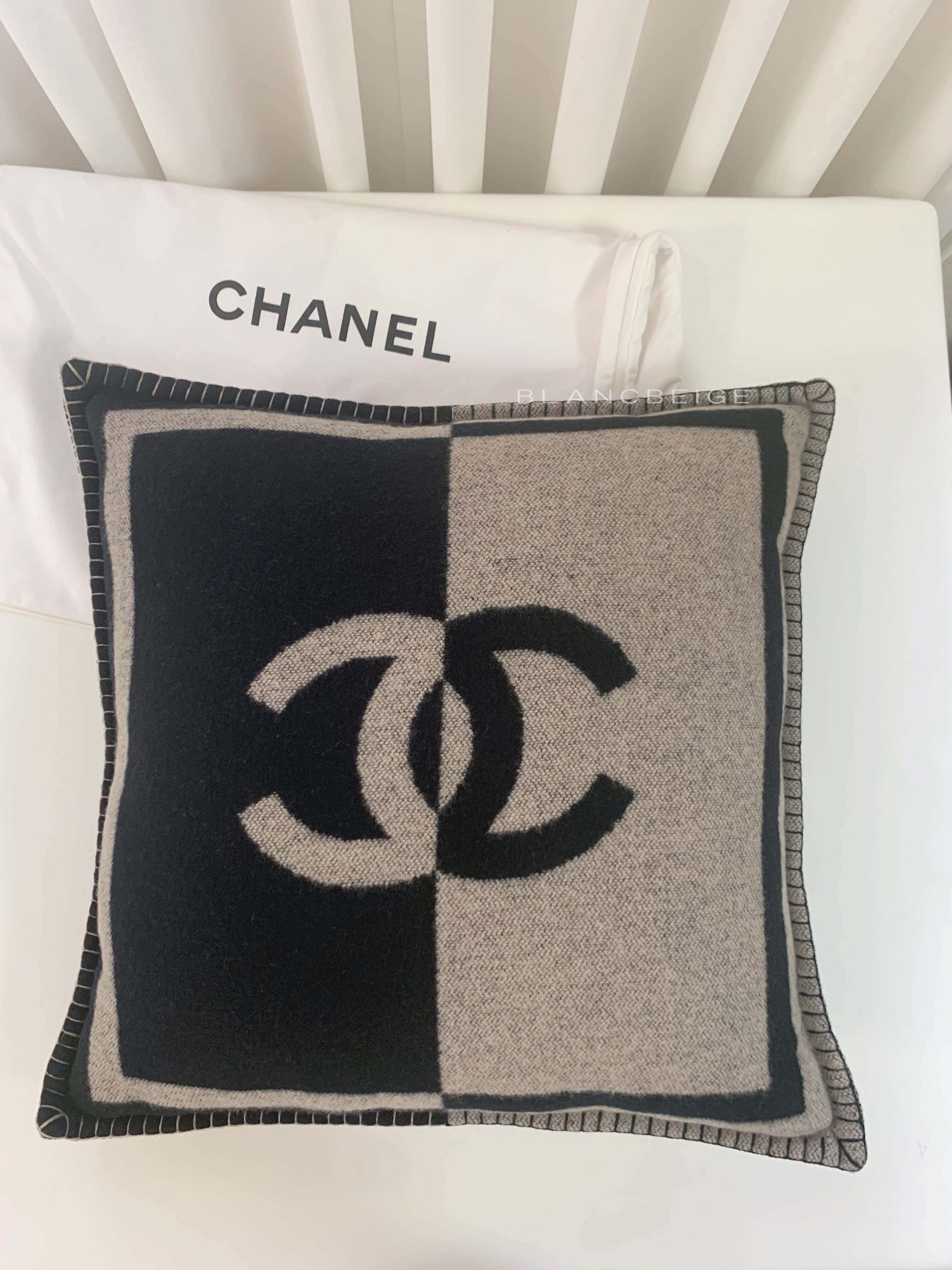 經典香奈兒標誌 壁紙 — 一個黑色和米色的 Chanel 標誌枕頭放在一個帶有 'CHANEL' 文字的白色枕頭上，背景是白色板條床頭板。