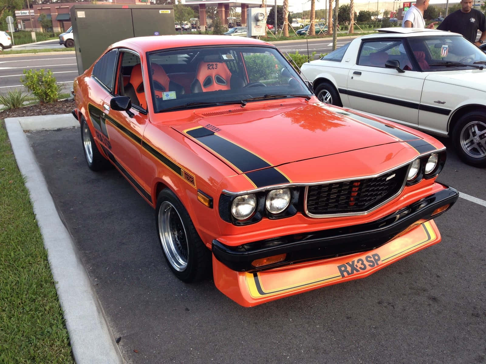 經典馬自達RX-3巔峰時刻 壁紙 — 一輛充滿活力的橙色經典RX3 SP跑車，帶有黑色賽車條紋，在黃昏時停放在停車場。