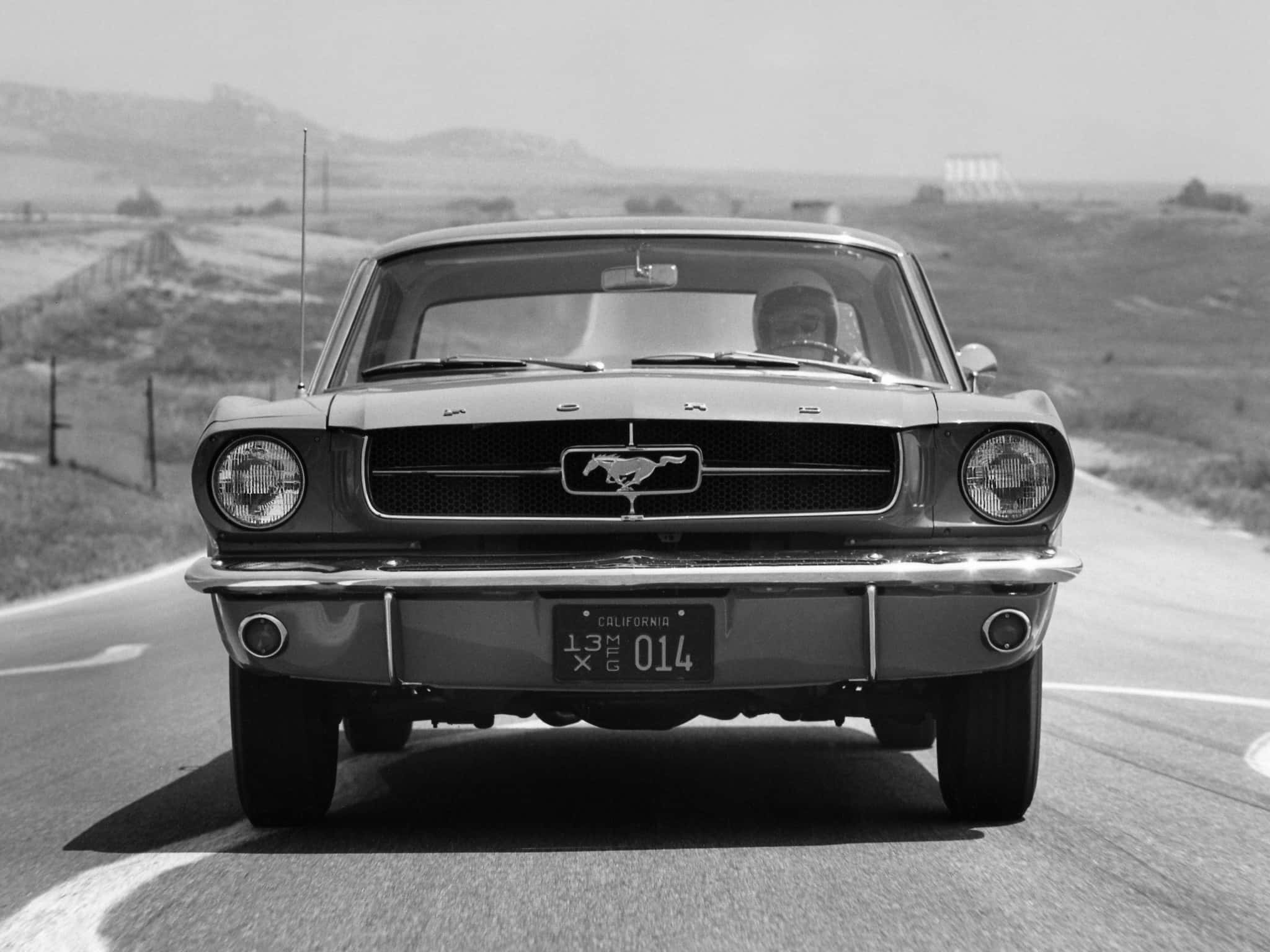 經典魅力 - 經典福特Mustang 壁紙 — 一輛經典福特野馬在鄉村道路上行駛，背景是丘陵，一張引人注目的黑白照片。