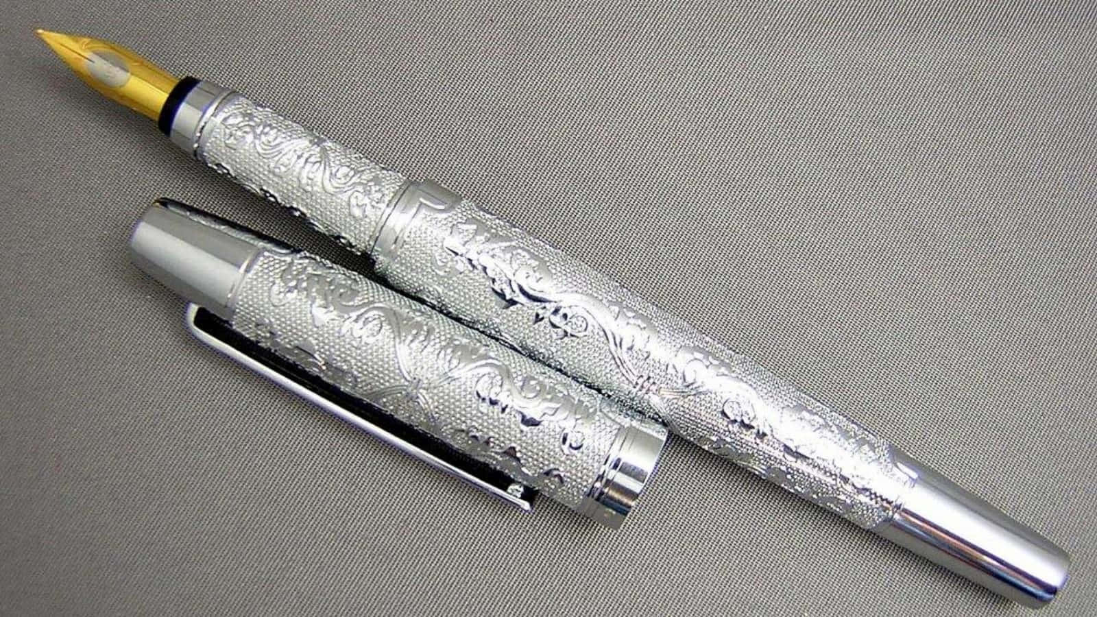 經典黑筆，準備留下印記 壁紙 — 一支銀色鋼筆，帶有精緻的花卉雕刻和金色筆尖，放在有紋理的灰色布料上，旁邊是筆蓋。