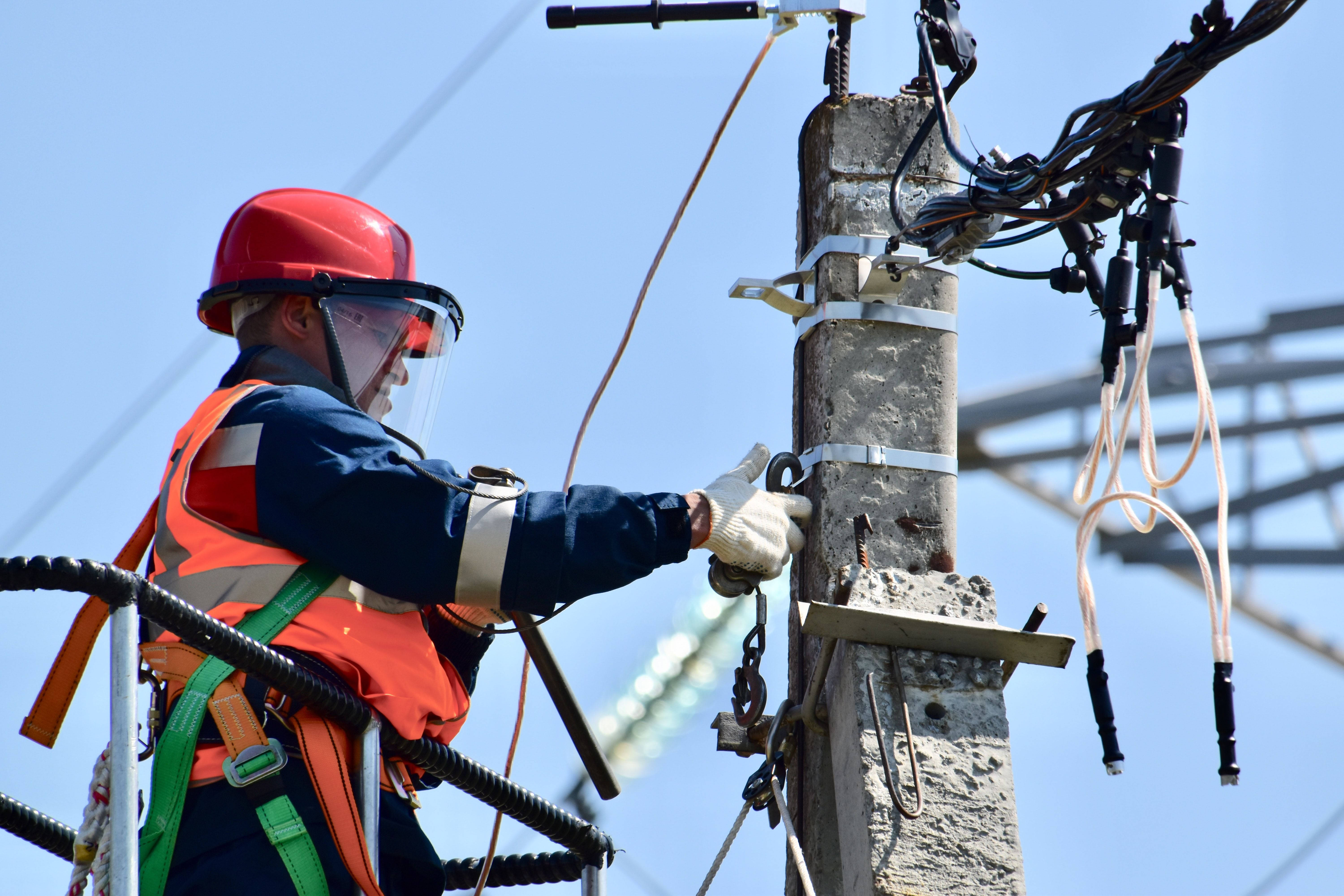 經驗豐富的電工修復電線 壁紙 — 電工戴著紅色頭盔和橙色背心，在晴朗的藍天下，在風化的混凝土電線桿上工作。