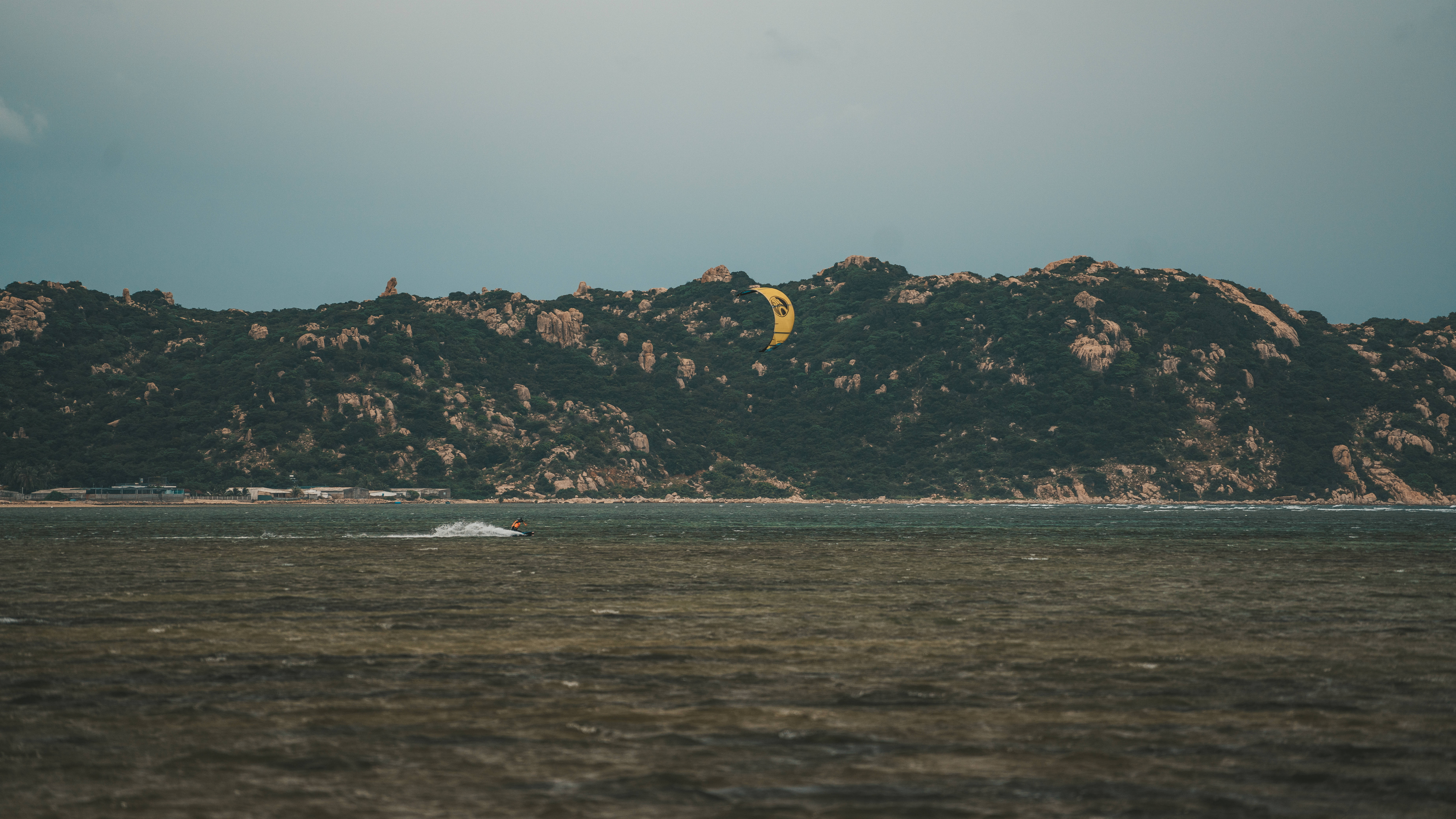 綠島附近的風帆衝浪 壁紙 — 風箏衝浪者在平靜的水面上滑行，明亮的黃色風箏在陰天之下飛越崎嶇的綠色山丘。