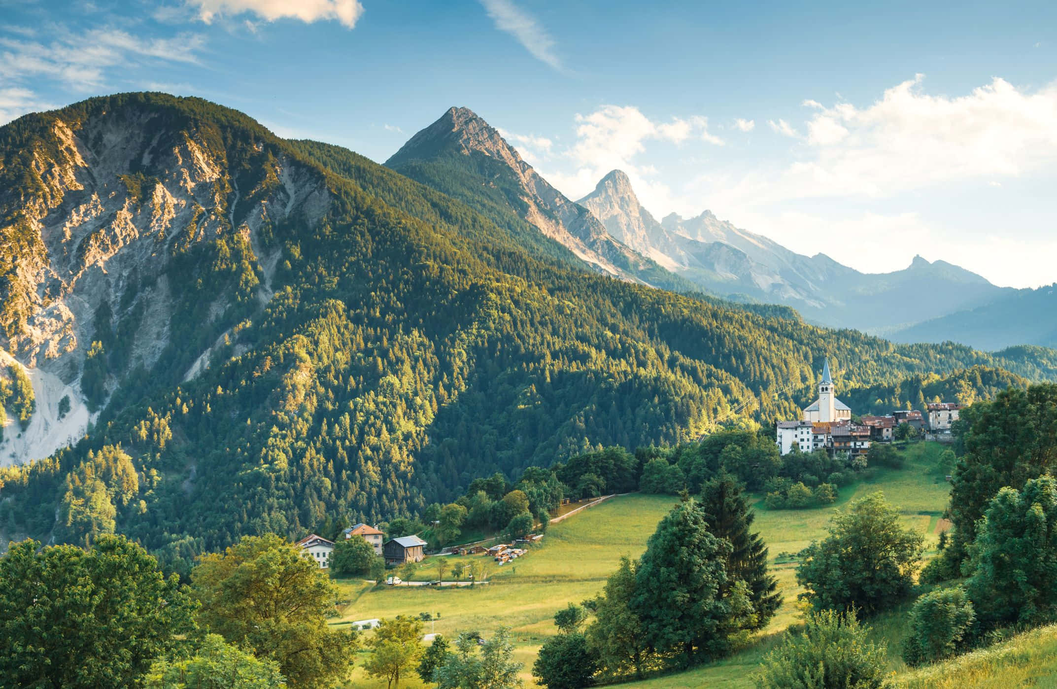 綠意盎然的山林美景 壁紙 — 一個陽光普照的阿爾卑斯山村，坐落在鬱鬱蔥蔥的綠色山谷中，高聳的森林覆蓋的山脈在明亮的藍天下。