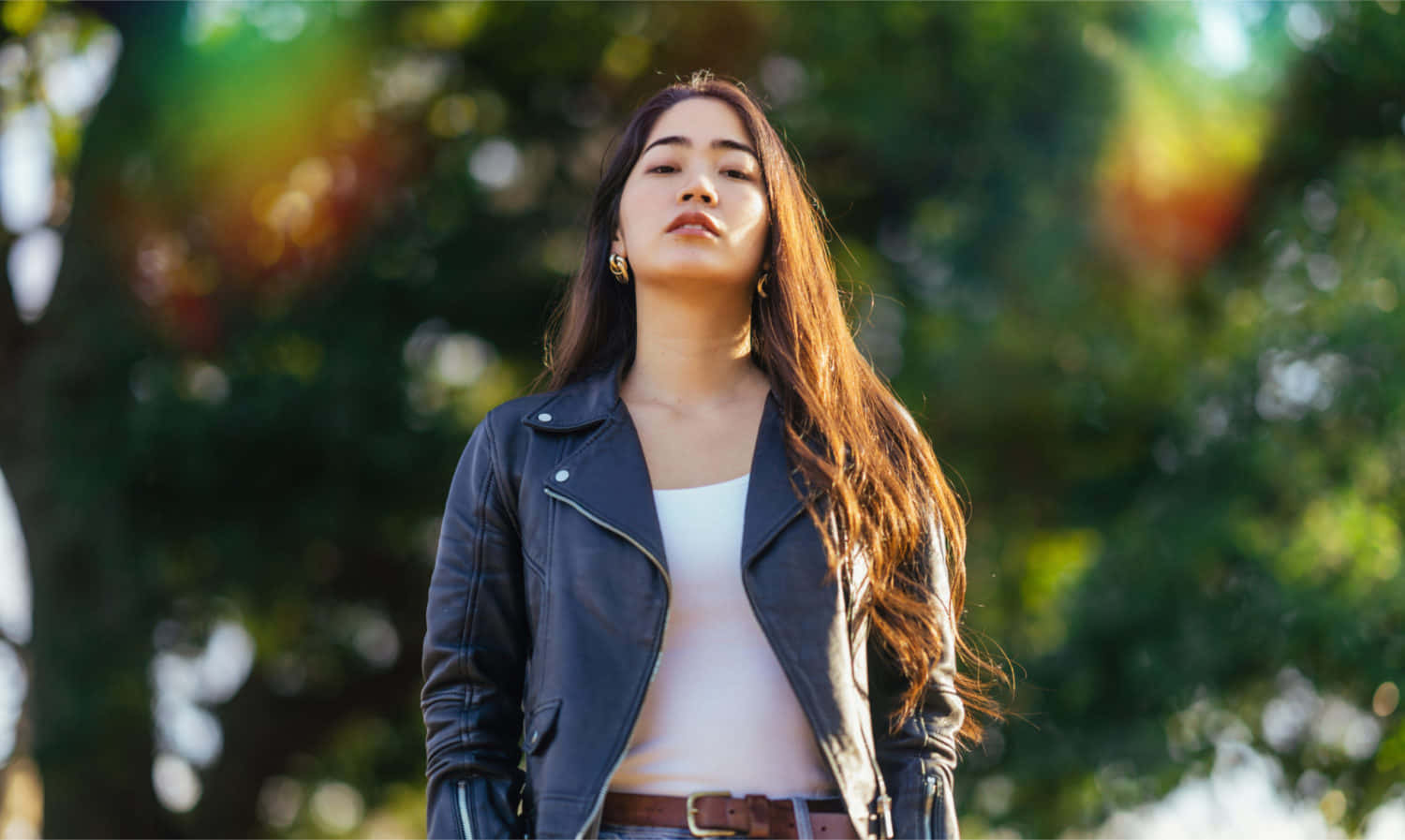綠樹模糊縮放背景 壁紙 — 一位穿著黑色皮褸的年輕女性站在戶外，閉上眼睛，享受透過綠樹灑落的溫暖陽光。
