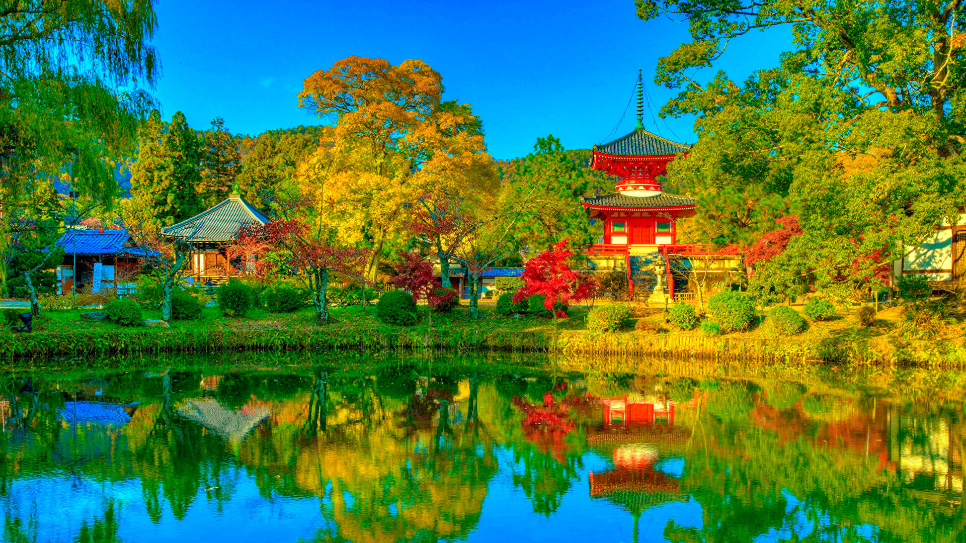 綠湖 日本風景 壁紙 — 鮮豔的紅色寶塔和傳統建築倒映在寧靜的池塘中，周圍是秋天的樹木，天空清澈湛藍。