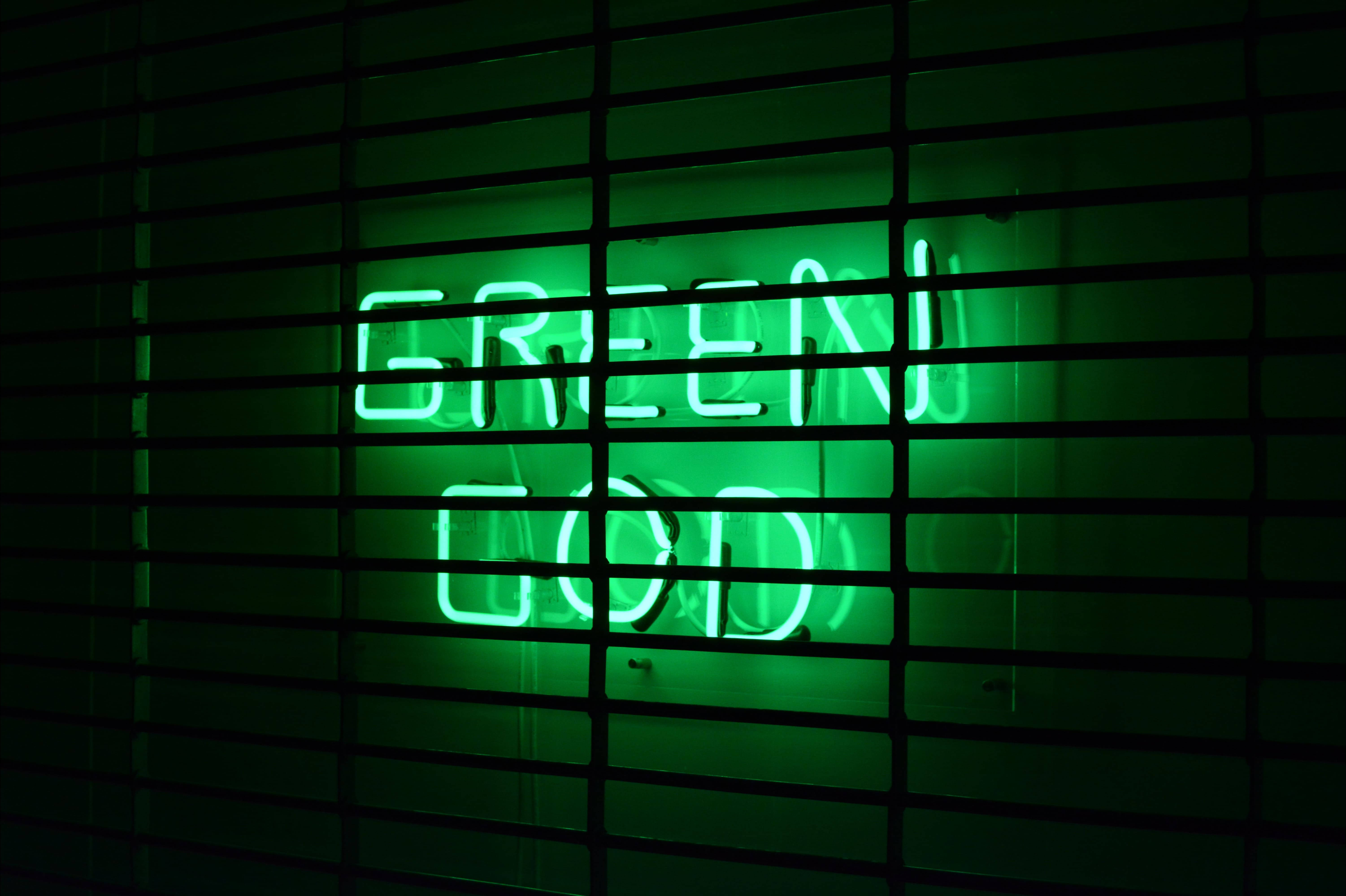 綠神霓虹綠招牌 壁紙 — 一個發光的綠色霓虹燈牌，上面寫著 'GREEN GOD'，透過暗色水平百葉窗在昏暗的房間中看到。
