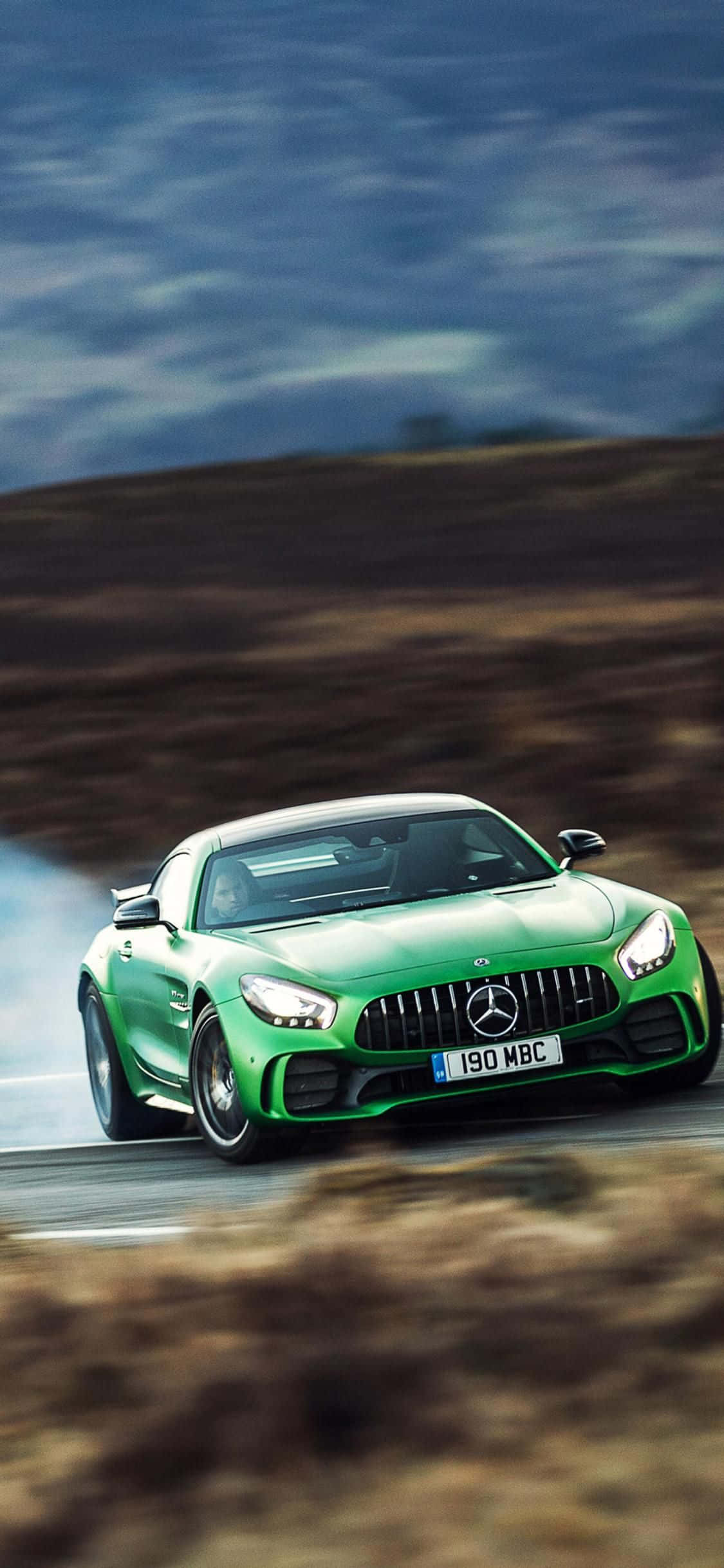 綠色 GT iPhone Xs Mercedes AMG 背景 壁紙 — 一輛充滿活力的綠色Mercedes-AMG GT R在蜿蜒的道路上飄移，伴隨著動態模糊和輪胎煙霧，背景是陰天。