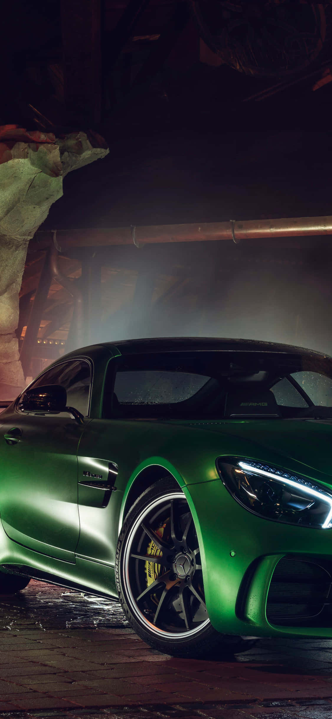 綠色 iPhone XS Mercedes AMG 夜間背景 壁紙 — 一輛時尚的啞光綠色跑車在骯髒的工業車庫中，在戲劇性的燈光下閃耀。