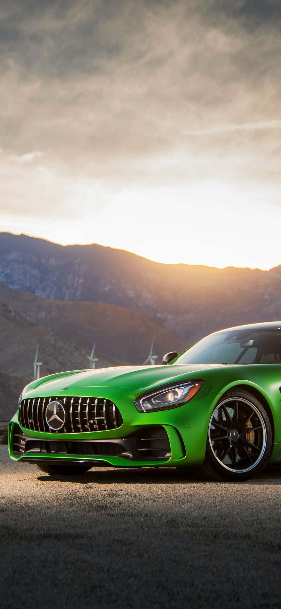 綠色AMG GT-R日出山景壁紙 壁紙 — 一架充滿活力的綠色 Mercedes-AMG GT 跑車停在瀝青路上，背景有風力渦輪機和山脈，天空呈現金色日落。