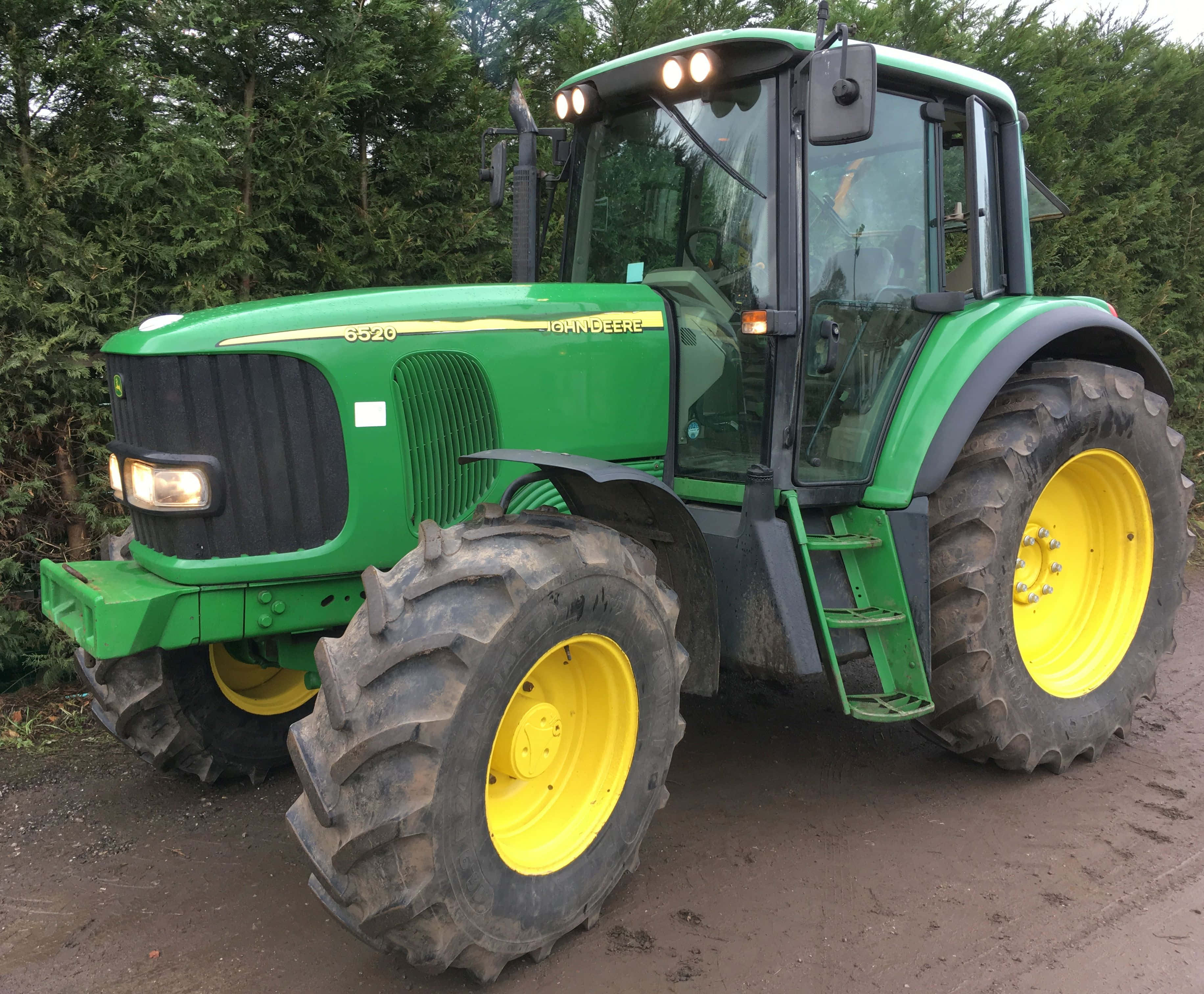 綠色John Deere 6920s拖拉機 壁紙 — 一部綠色和黃色的John Deere 6520拖拉機，帶有大型泥濘輪胎，停放在常綠樹附近的泥土上。