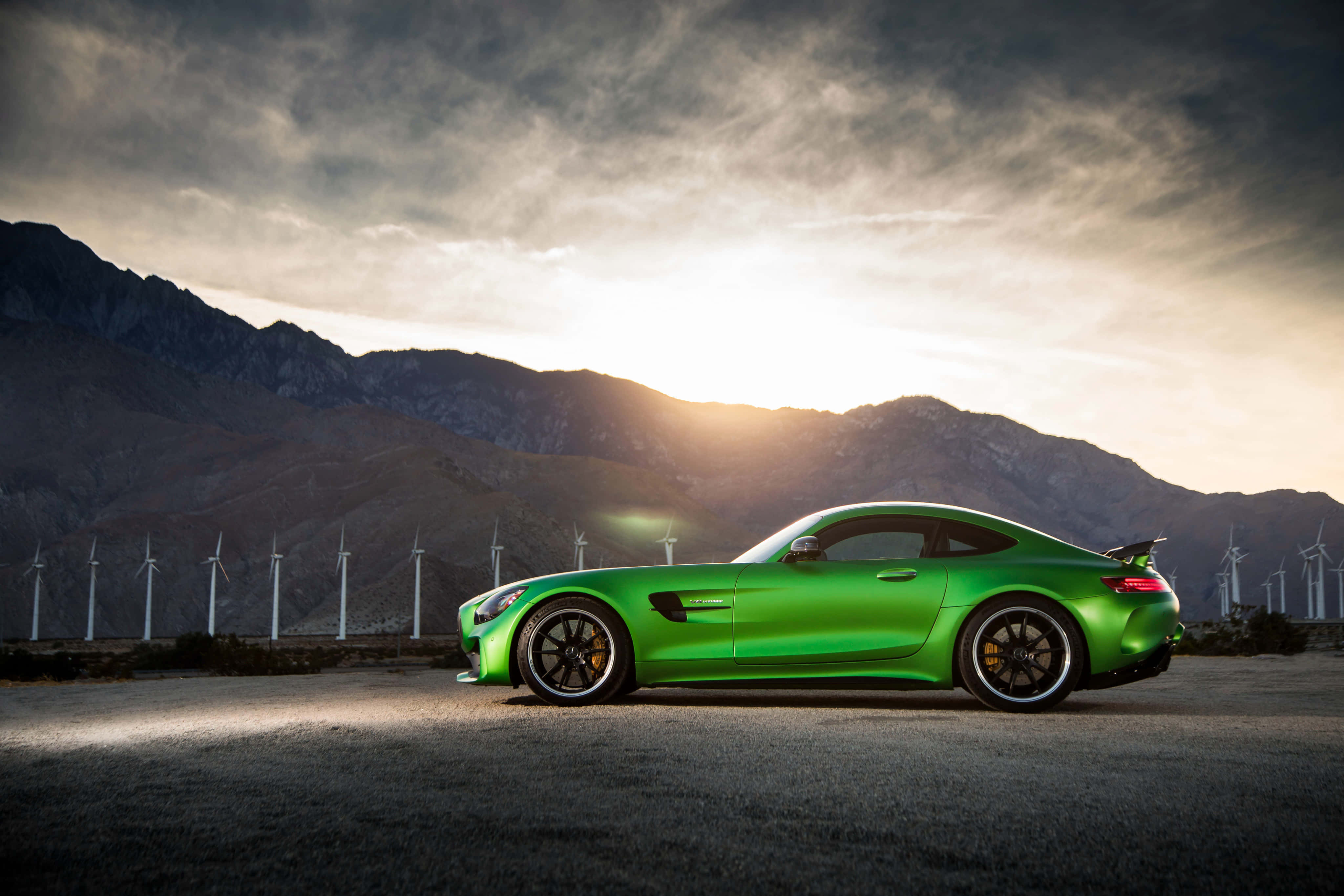 綠色梅賽德斯-AMG GT R 與夕陽山景背景 壁紙 — 一輛充滿活力的綠色跑車在金色夕陽下閃閃發光，背景是山脈和風力發電機。