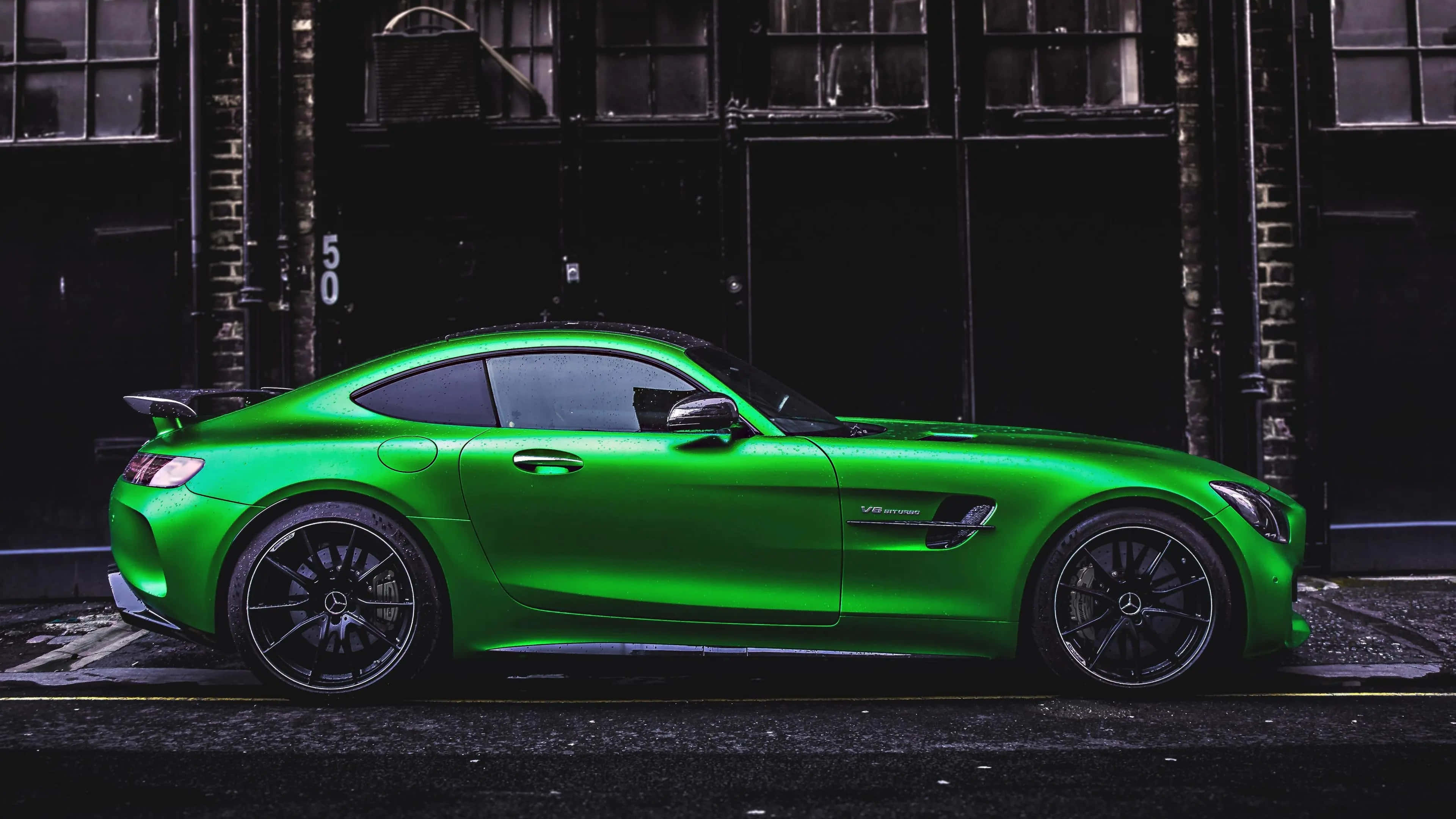 綠色梅賽德斯 AMG GT R 側面角度背景 壁紙 — 一輛充滿活力的綠色 Mercedes-AMG GT 跑車停放在一條光線昏暗的都市小巷中，小巷有磚牆和工業窗戶。
