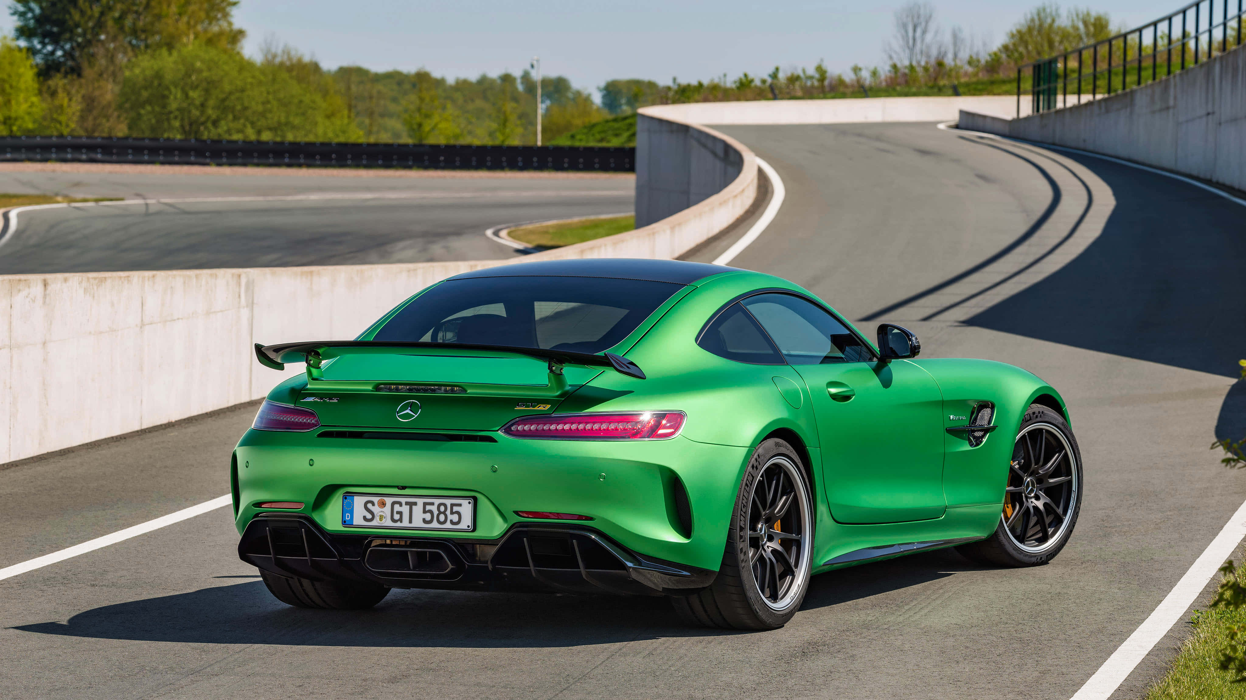 綠色梅賽德斯-AMG GT R車尾角度背景 壁紙 — 一輛充滿活力的綠色 Mercedes-AMG GT R 跑車在晴朗天空下，沿著由混凝土護欄和綠樹環繞的彎曲瀝青賽道行駛。