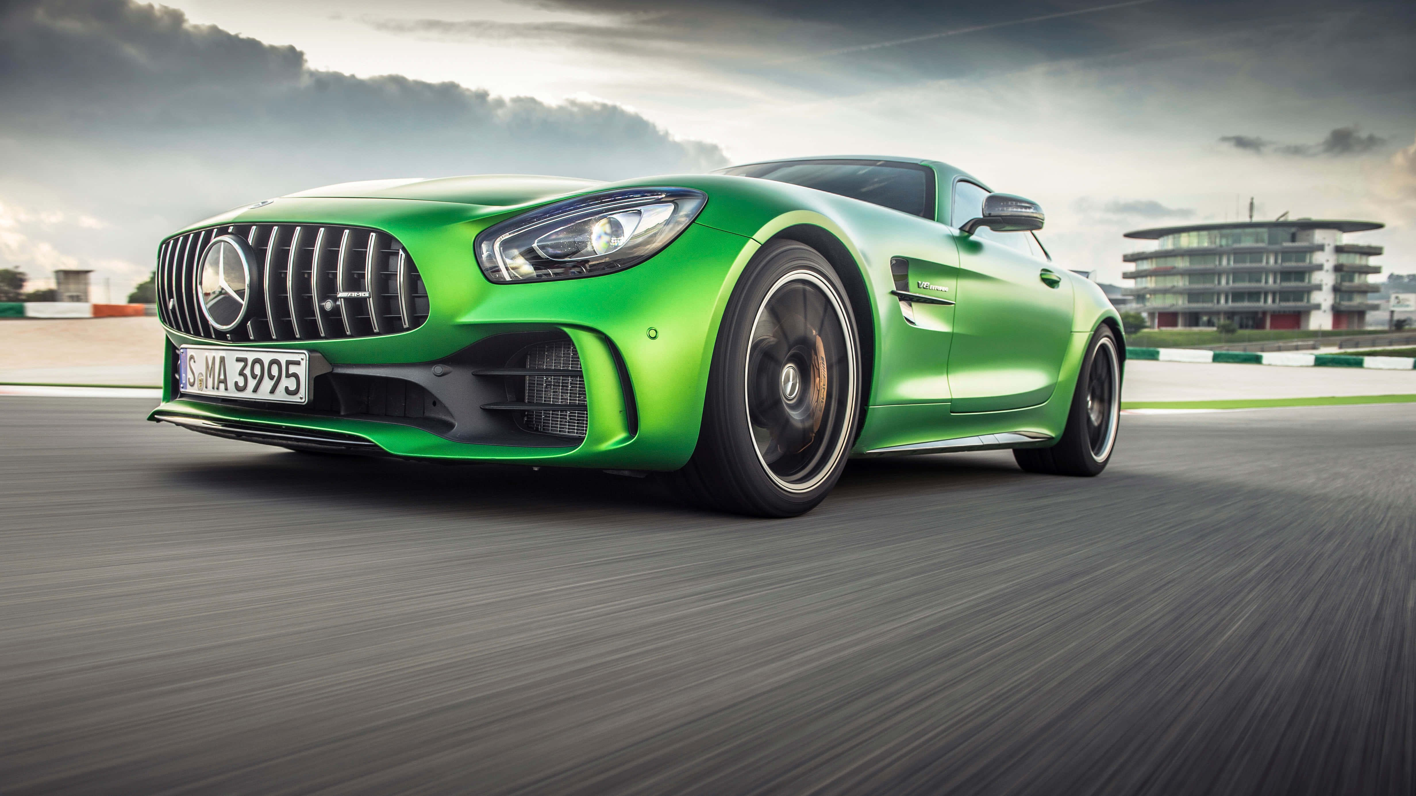 綠色梅賽德斯AMG GT R疾馳背景 壁紙 — 一輛充滿活力的綠色Mercedes-AMG GT S在陰天下的賽道上疾馳，瀝青路面呈現動態模糊效果。