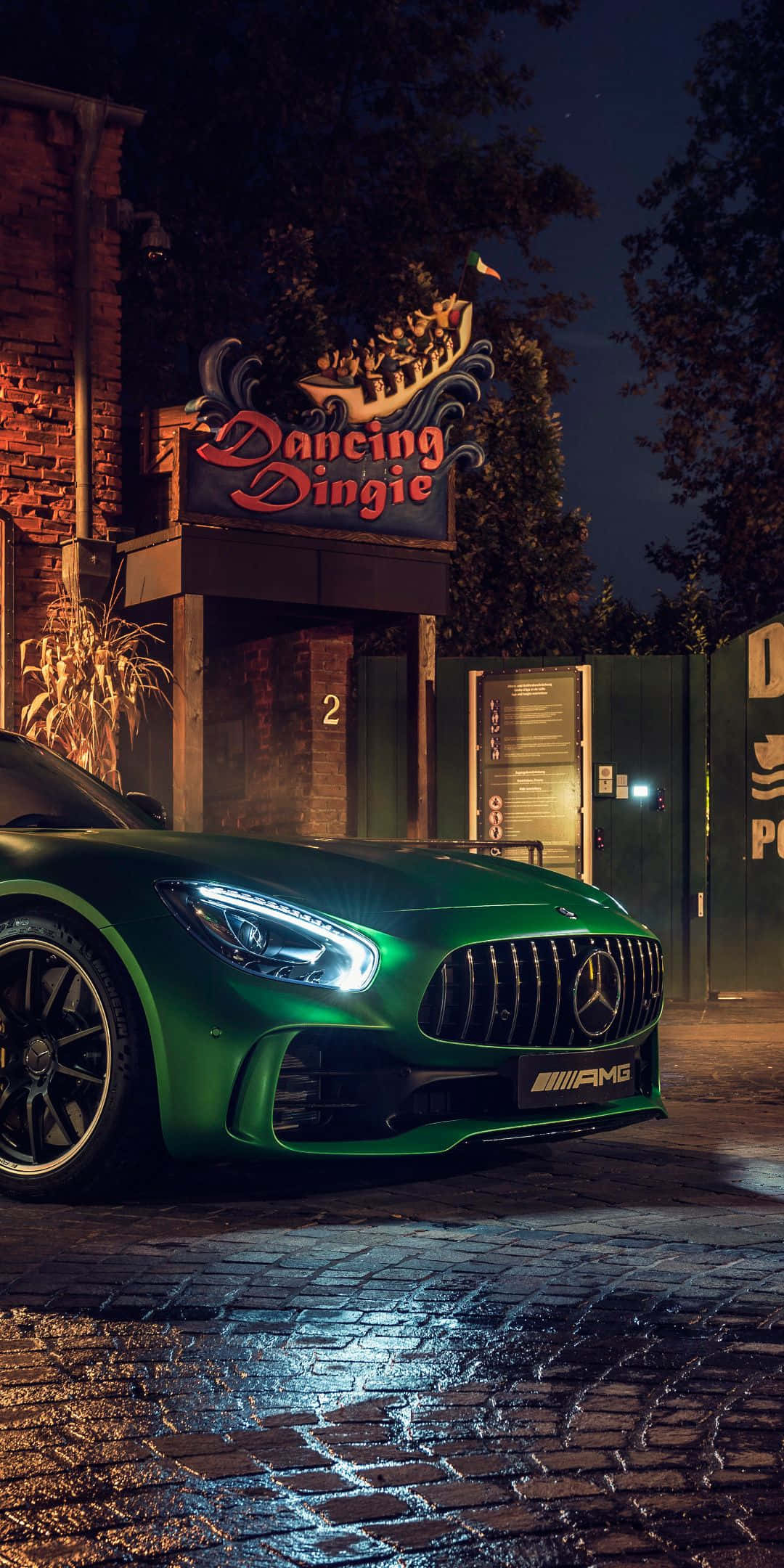 綠色梅賽德斯AMG GT R背景 壁紙 — 一輛充滿活力的綠色 Mercedes-AMG 跑車在夜間停泊在濕漉漉的鵝卵石街道上，靠近 'Dancing Dingle' 標誌，在霓虹燈下閃閃發光。