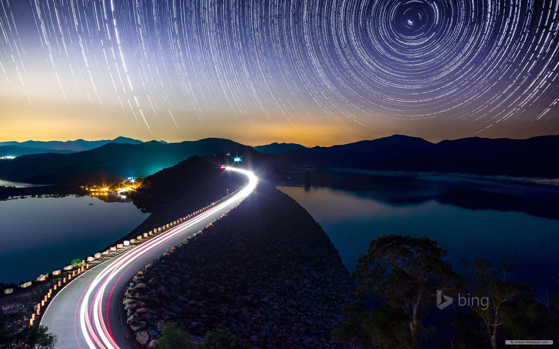 星軌在蜿蜒的道路上空盤旋，黃昏時水壩附近有汽車的光跡。