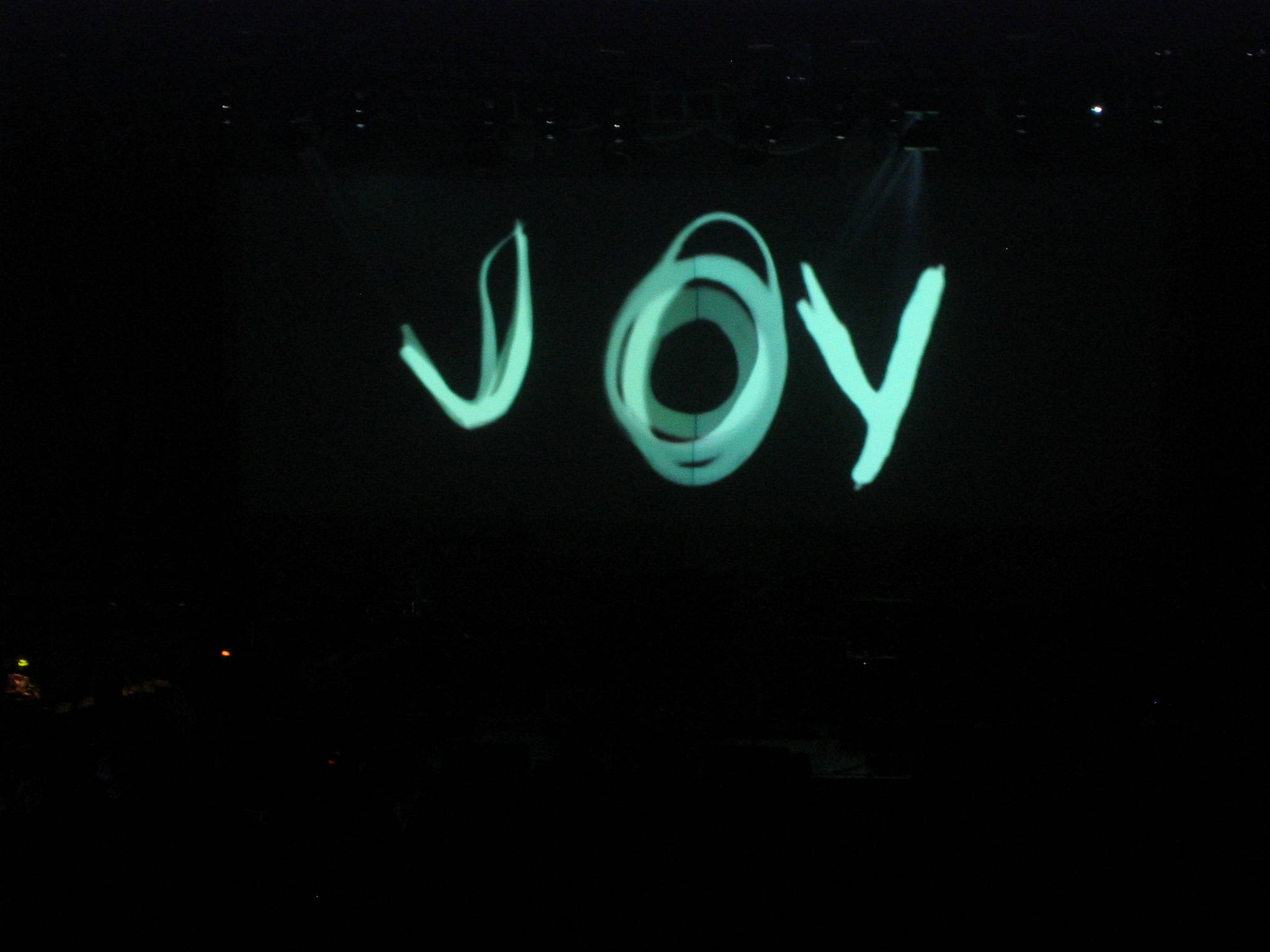 綠色歡樂塗鴉 壁紙 — 「JOY」這個詞在漆黑的舞台上散發著充滿活力的藍綠色光芒，在黑暗的音樂會場地被聚光燈照亮。