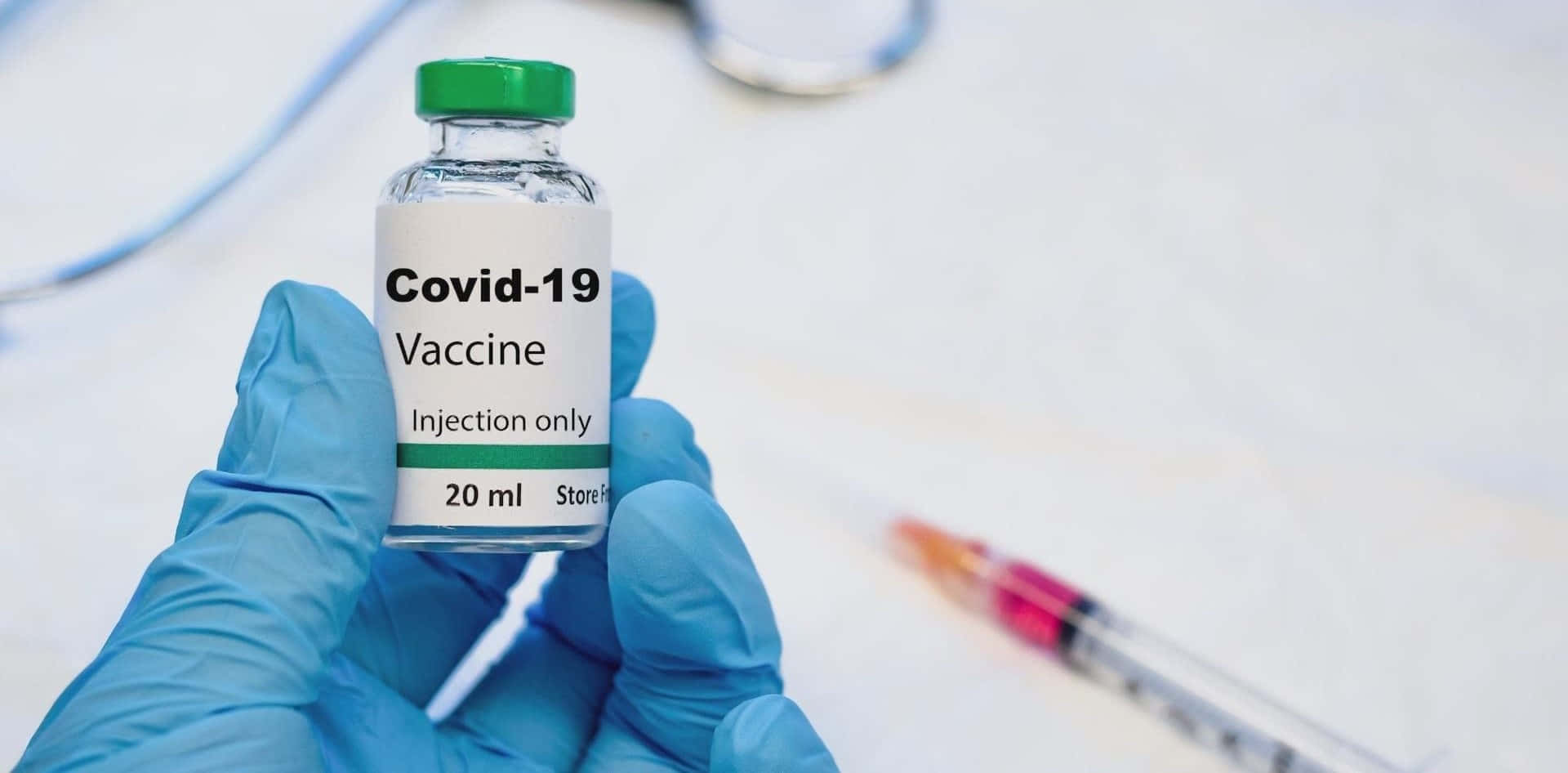 藍色醫用手套的手握住一個Covid-19疫苗小瓶，附近有注射器。