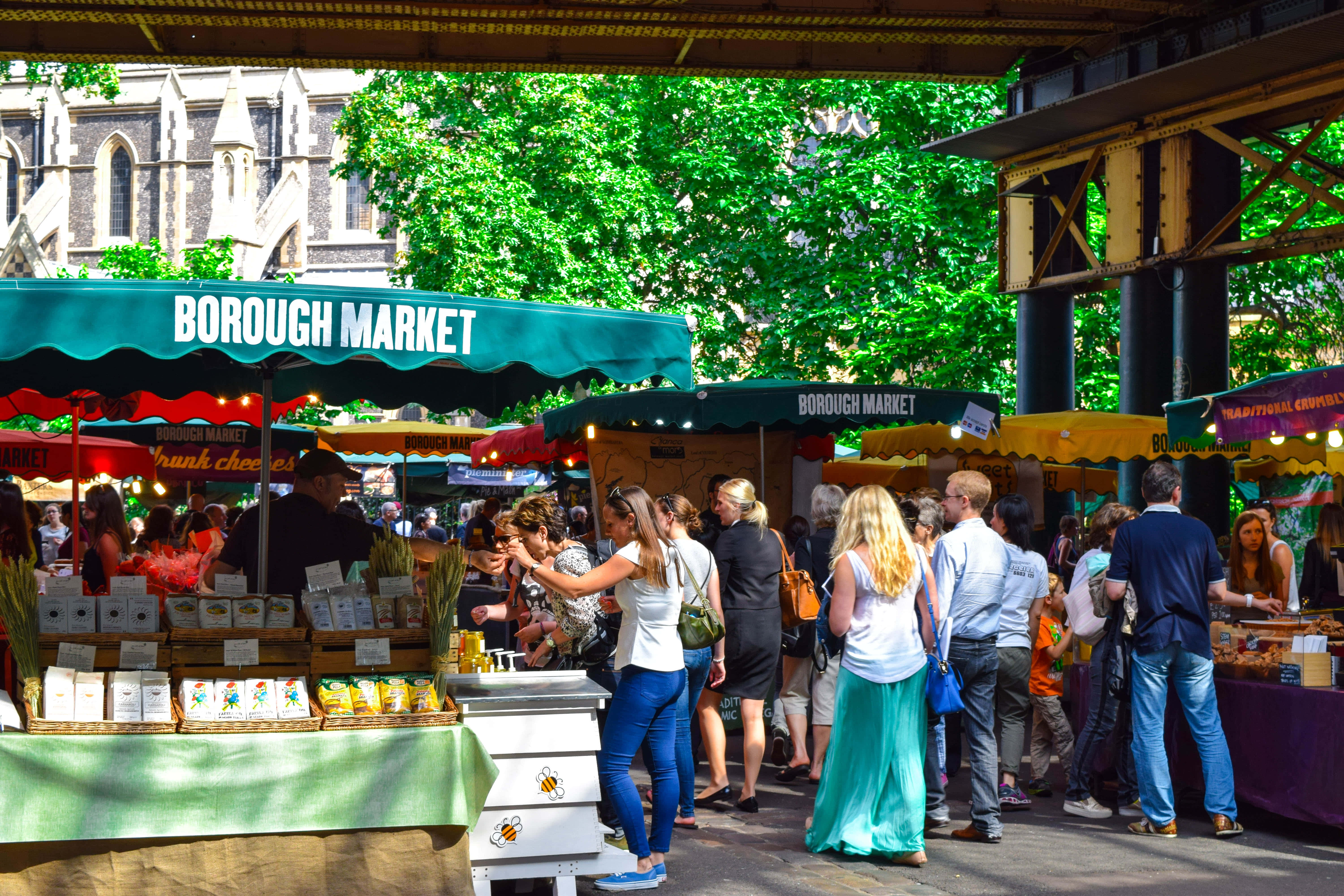 Farmers Market 桌布 壁紙 — 綠色簷篷下擠滿人的波羅市場攤檔，購物者在明媚夏日陽光下瀏覽食物。