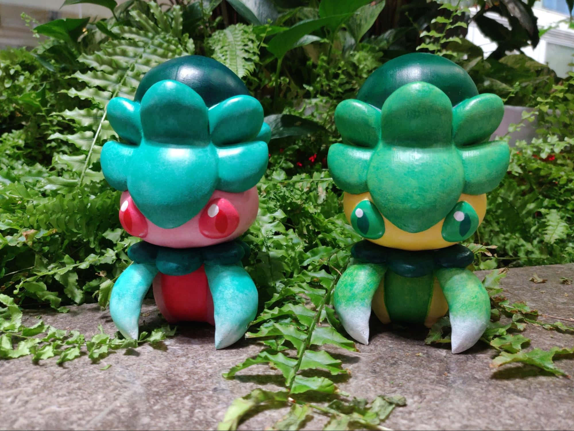 綠色與粉紅色寶可夢 Fomantis 壁紙 — 兩個手繪嘅 Bulbasaur 公仔並排坐喺石頭上，周圍係茂密嘅蕨類植物，喺花園環境入面。