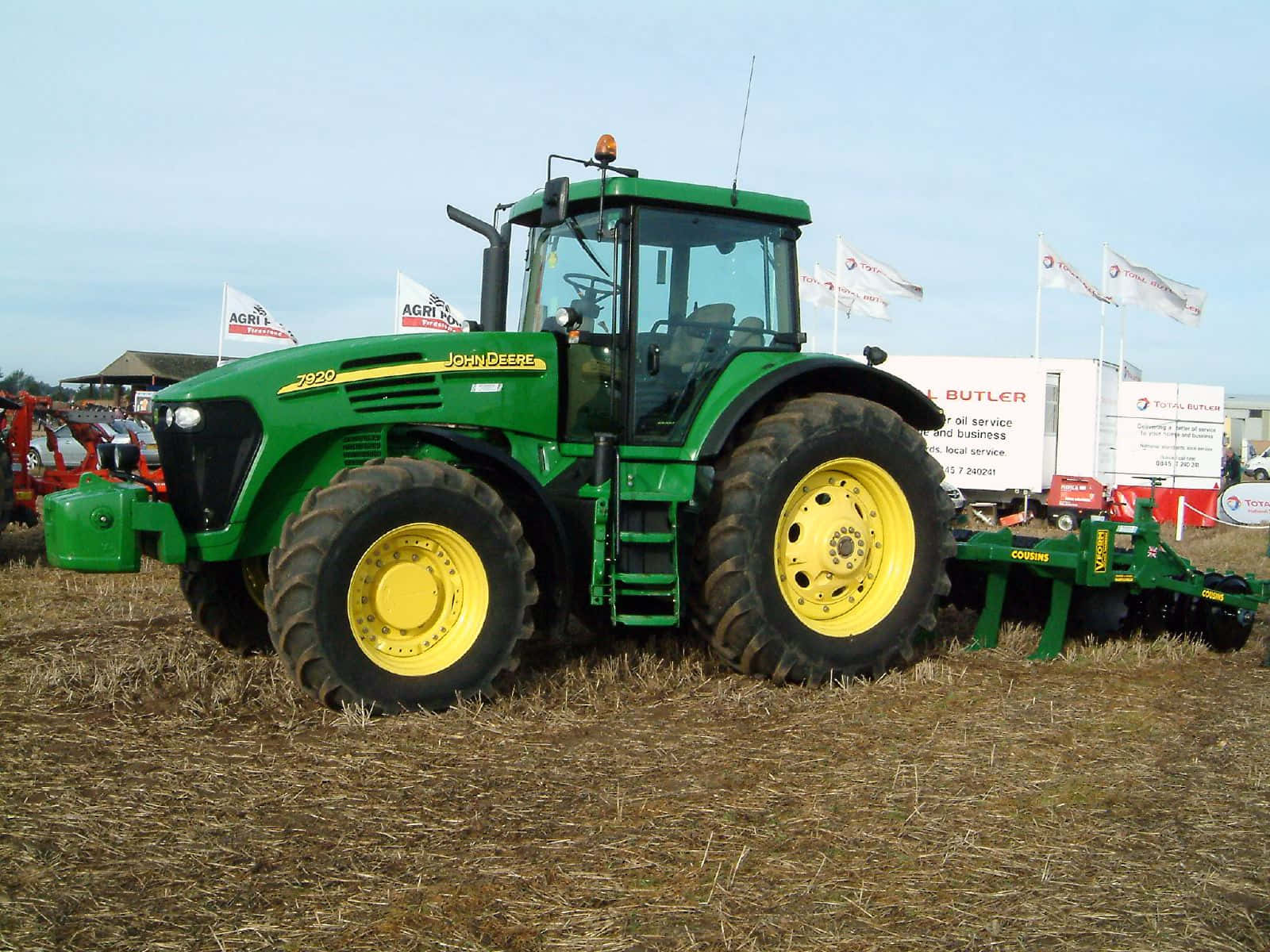 綠色重型拖拉機田間風景 壁紙 — 一部綠色和黃色的John Deere 7920拖拉機站在乾燥的殘茬田上，背景有農業設備和橫幅，天空晴朗。