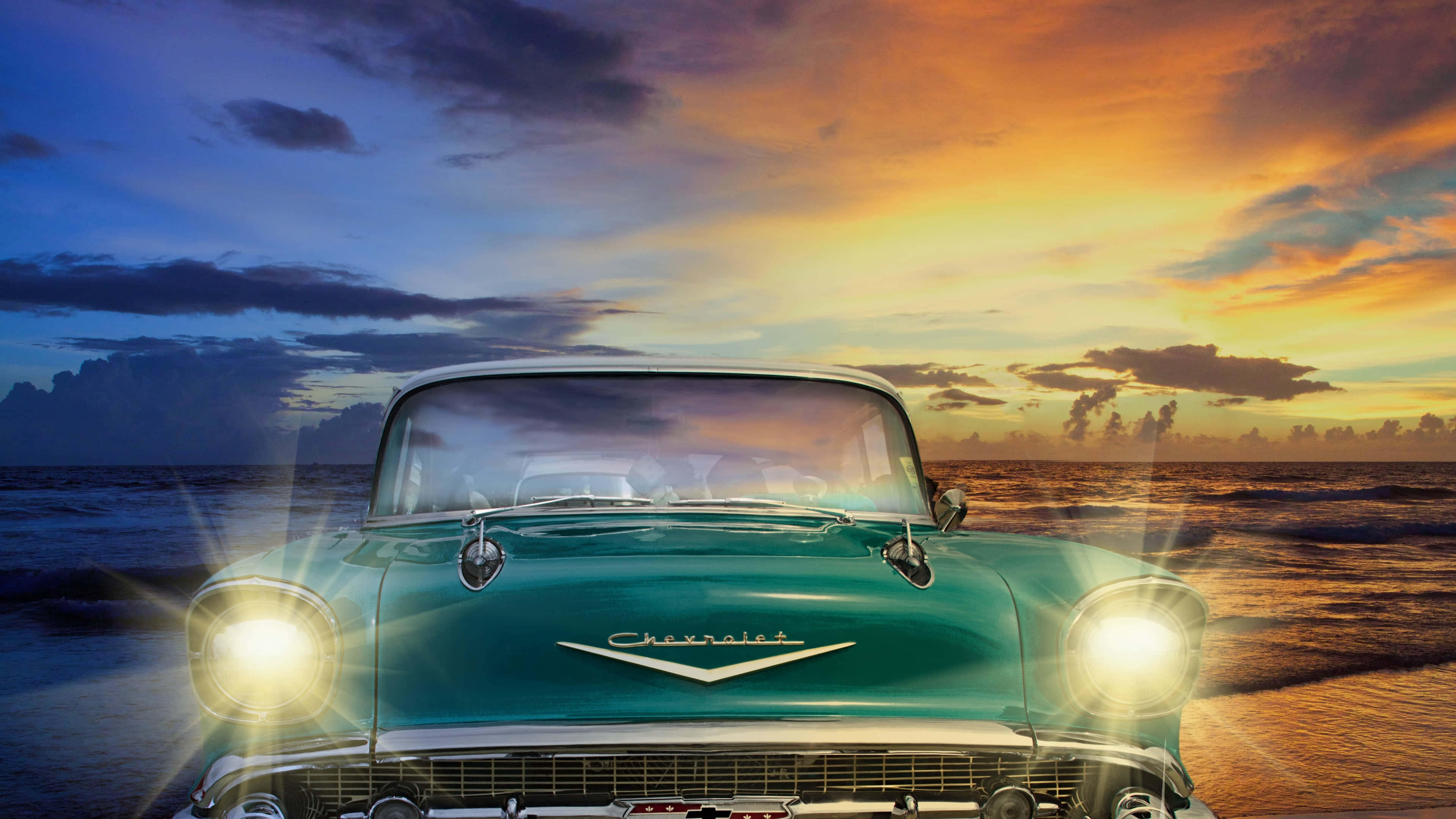 綠色雪佛蘭Bel Air經典車款 壁紙 — 一輛青綠色的1957年雪佛蘭，車頭燈發光，在沙灘上夕陽西下時面向觀眾，天空呈現鮮豔的橙色和藍色。