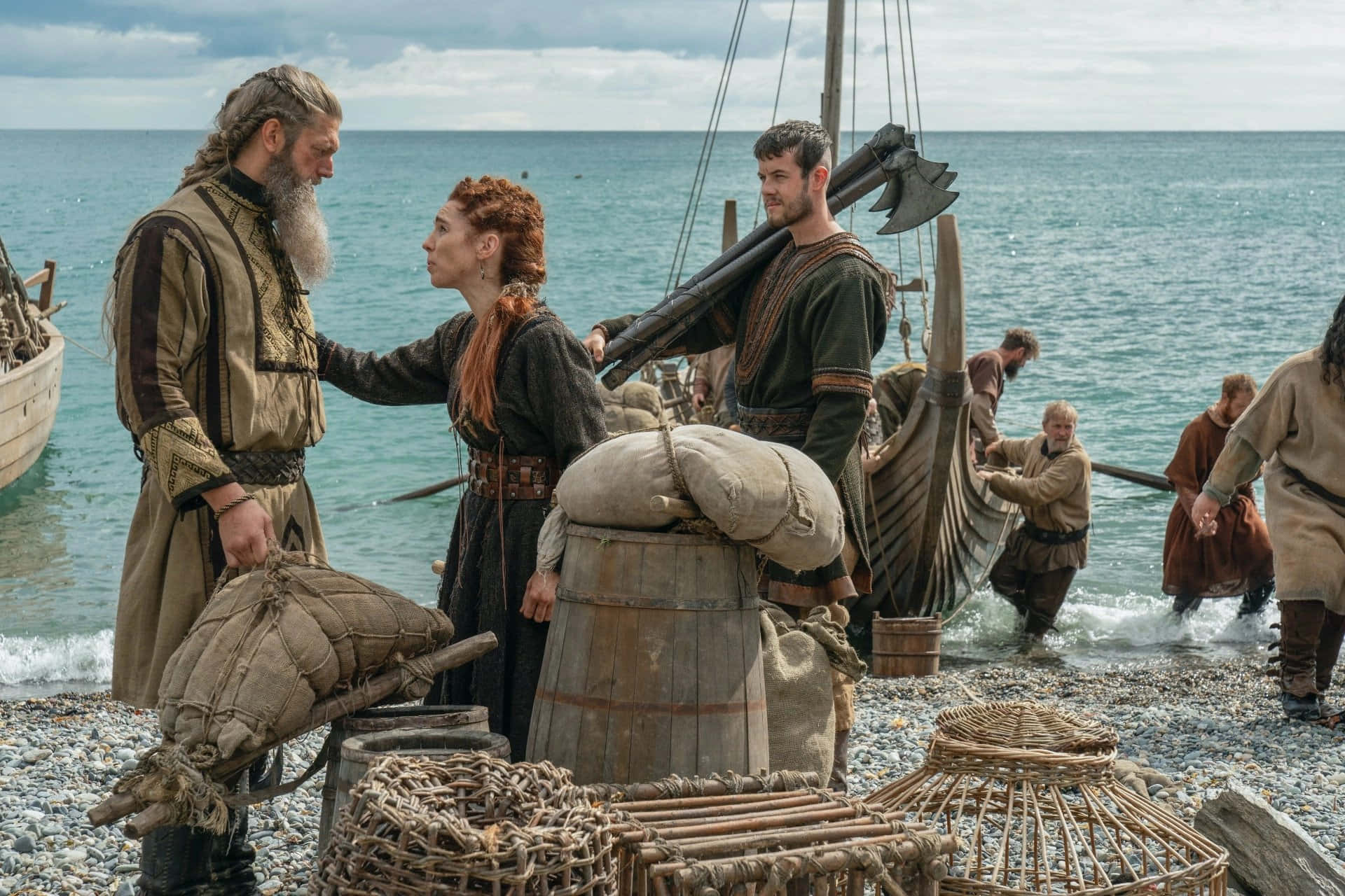 Vikings 桌布 壁紙 — 維京戰士和一個女人站在卵石灘上，旁邊是裝滿貨物的長船，天空多雲。