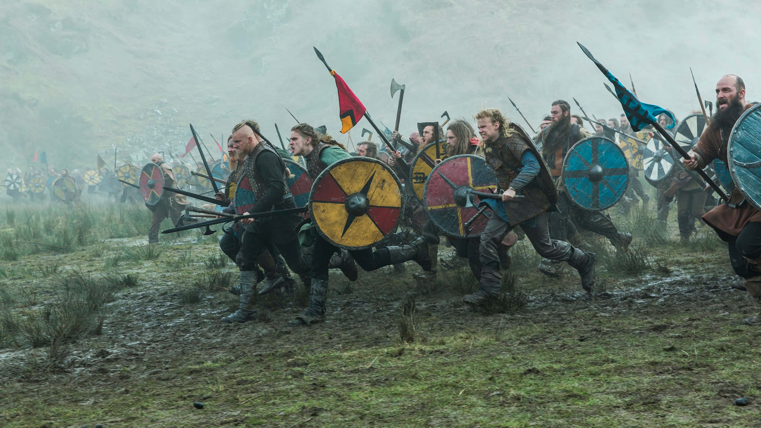 Vikings 桌布 壁紙 — 維京戰士在泥濘的田野中手持盾牌和長矛，在霧濛濛的灰色天空下向前衝鋒。