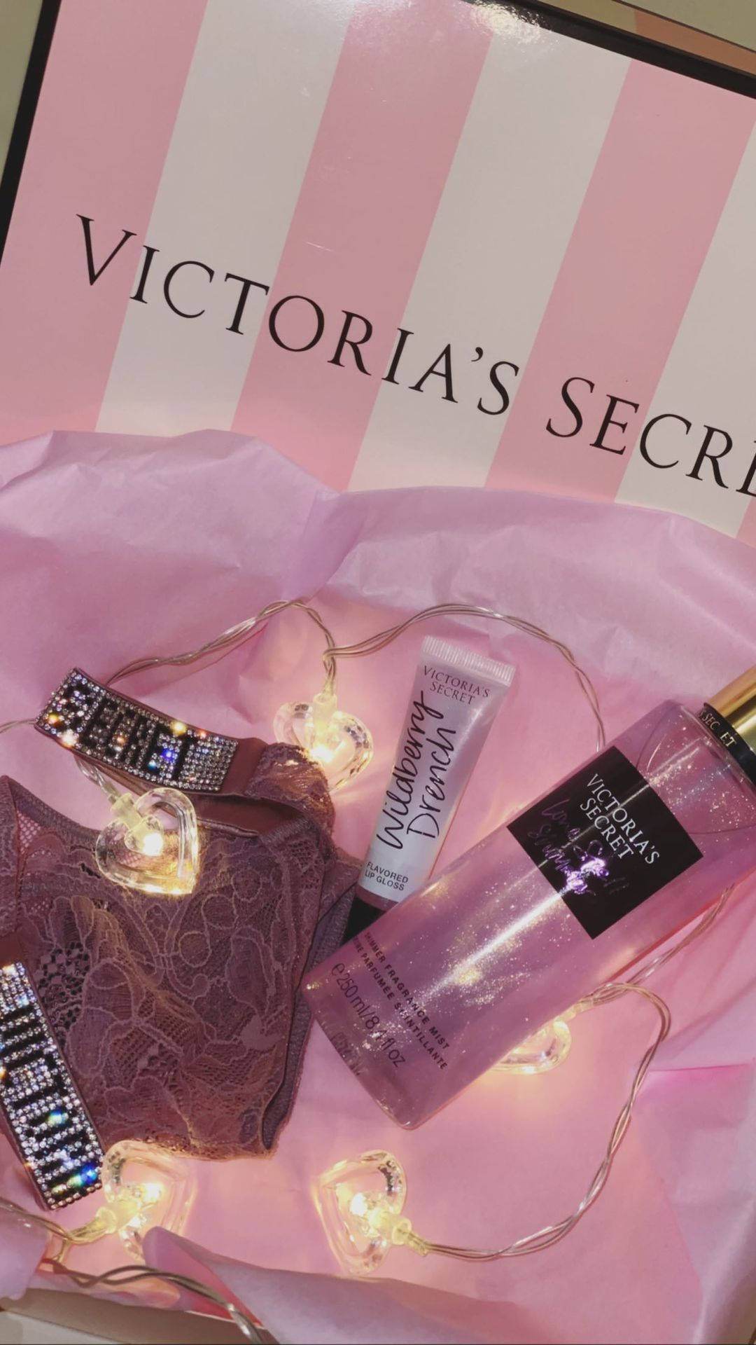 維多利亞的秘密禮品組合 壁紙 — 粉紅色Victoria's Secret內衣及身體產品在粉紅色紙巾上的串燈下發光。