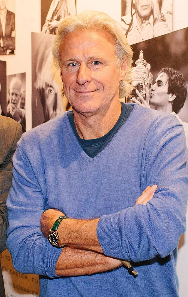 網球傳奇比約恩·博格比賽瞬間 壁紙 — 一位白髮藍眼的男士，身穿藍色毛衣，雙手交叉，在黑白照片的背景前自信地微笑。